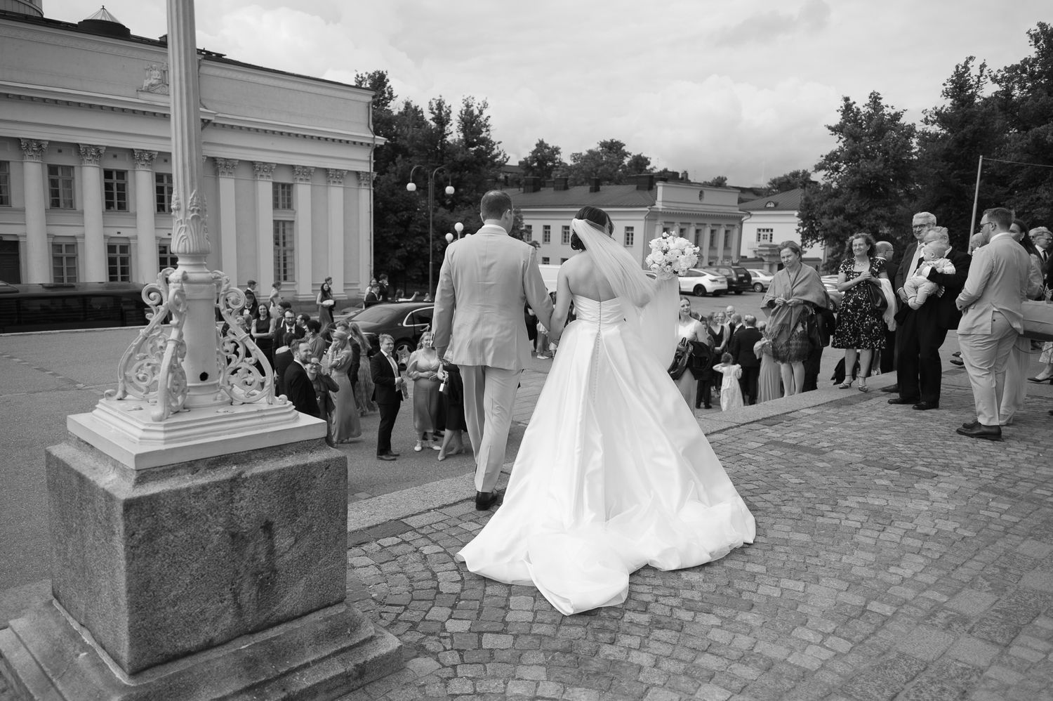 Black and white photo of a bride and groom walking toward guests outside a grand building at an outdoor wedding ceremony.