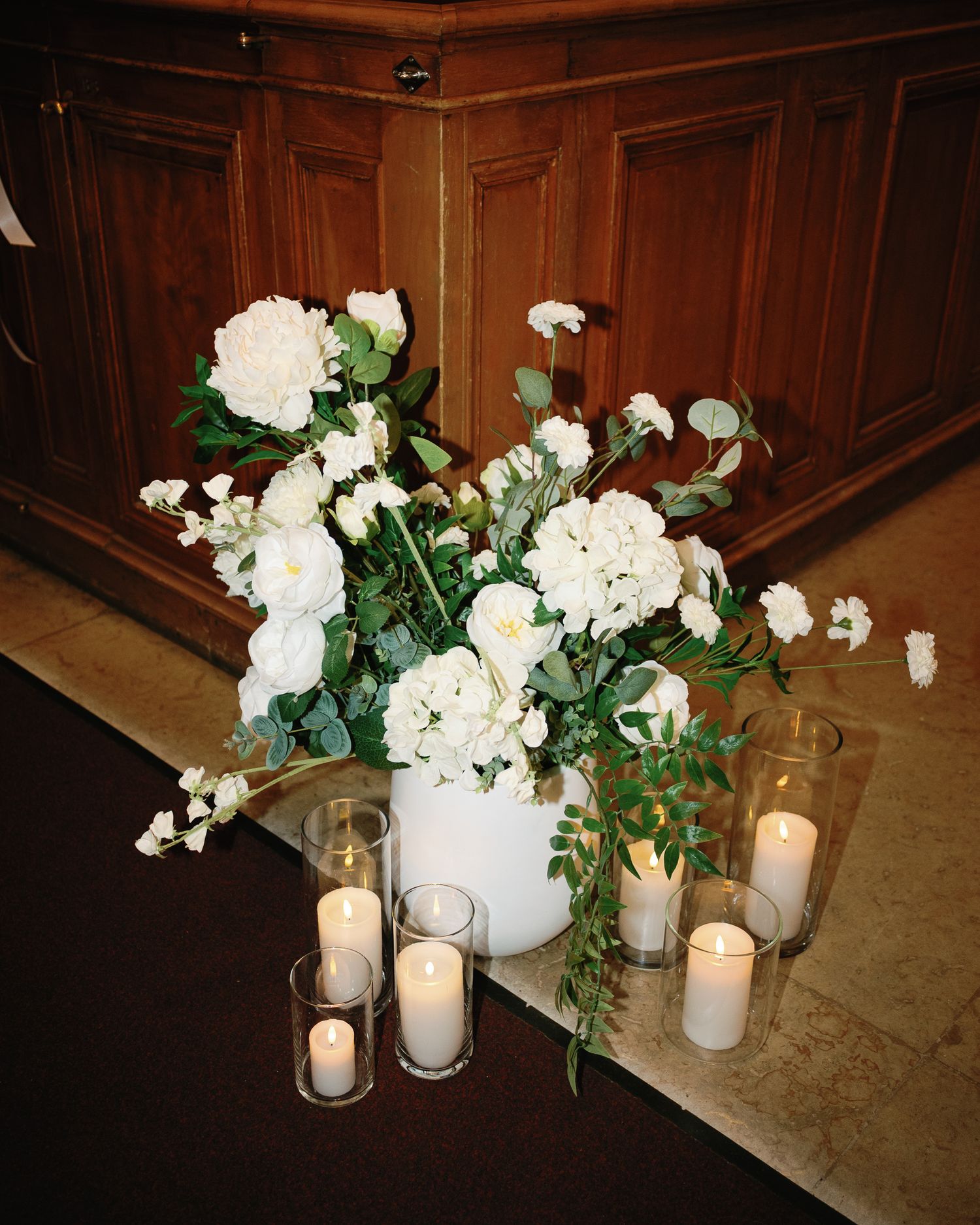 White floral arrangement with peonies and greenery in white vase surrounded by pillar candles on wooden floor.