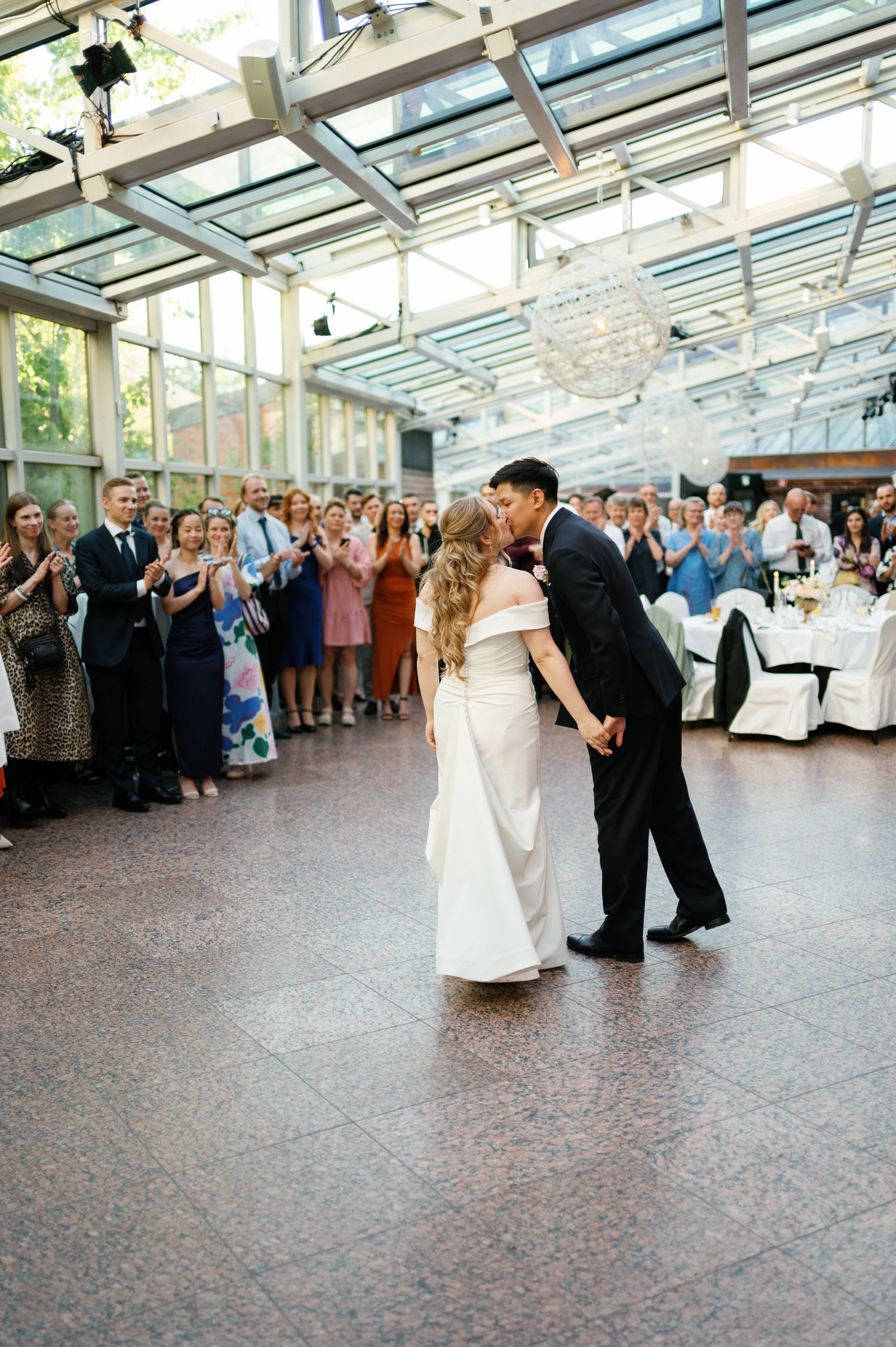 Bride and groom share first dance kiss in elegant glass-ceiling venue while wedding guests applaud around them.