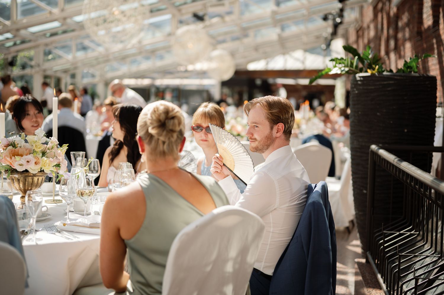 Guests smiling and chatting at an elegant wedding reception in a bright greenhouse venue with floral centerpieces.