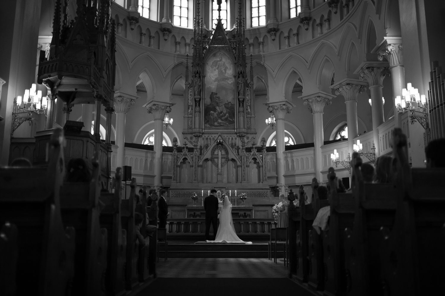 Black and white photo of a wedding ceremony at a grand church altar with bride and groom surrounded by guests.