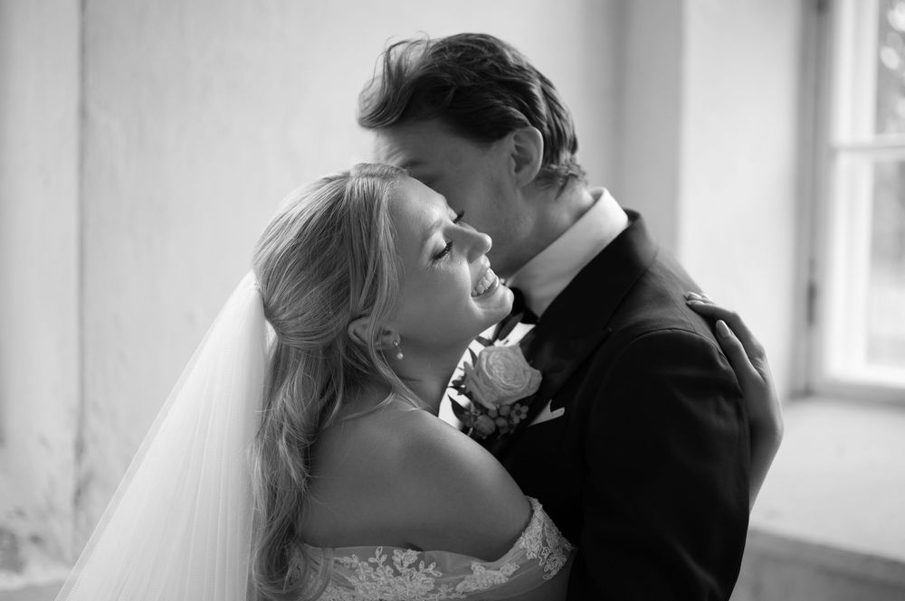 An artistic black and white wedding photo capturing a romantic embrace near a window.