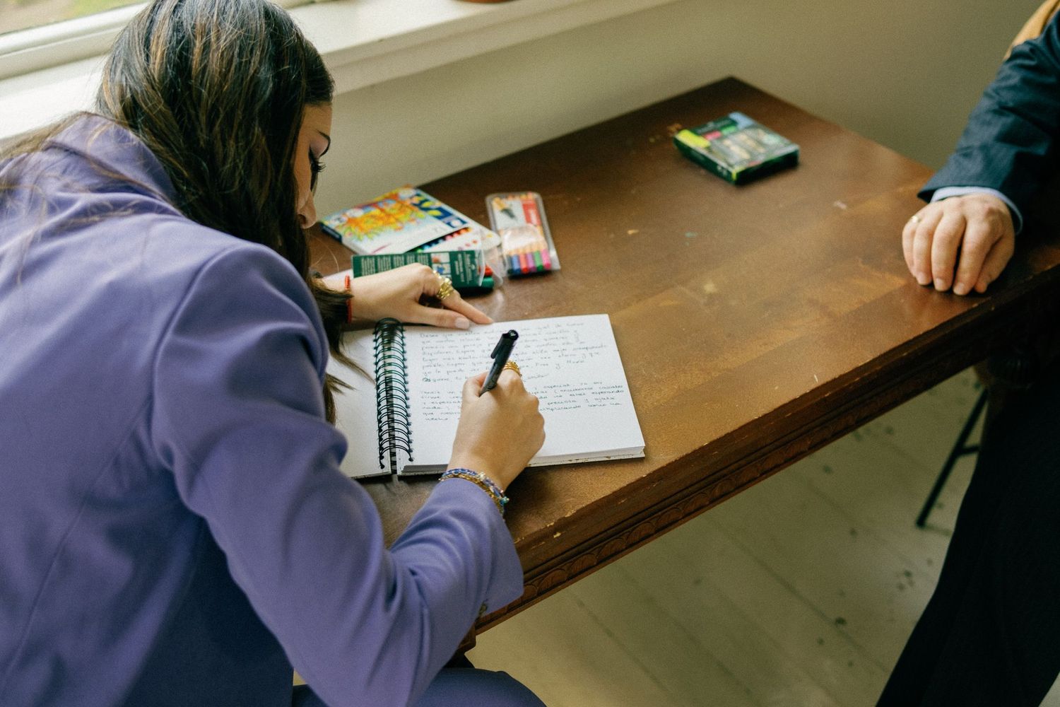People writing in notebooks while seated at a wooden table during an indoor event.