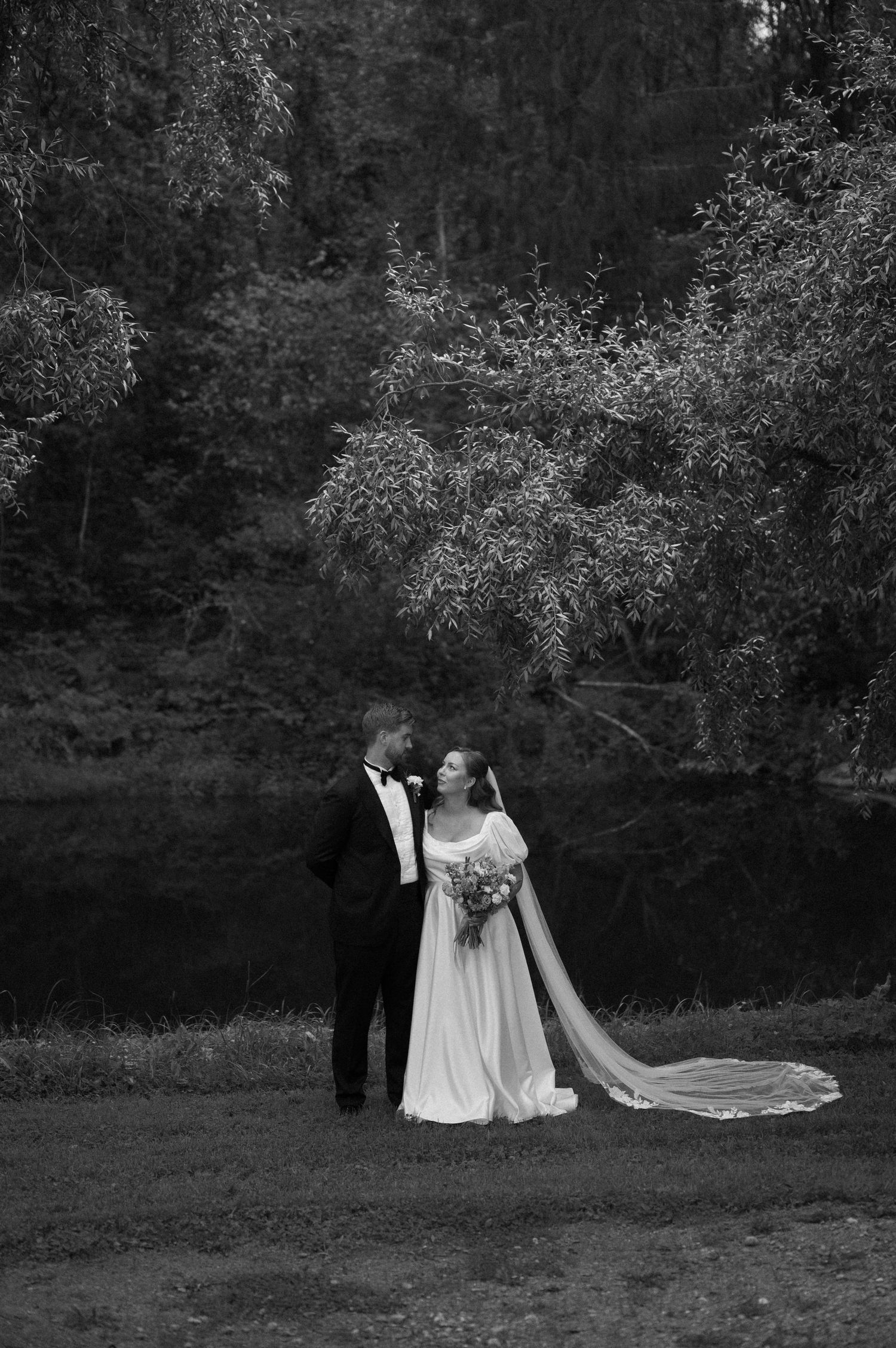 Black and white wedding photo of couple standing near water's edge.
