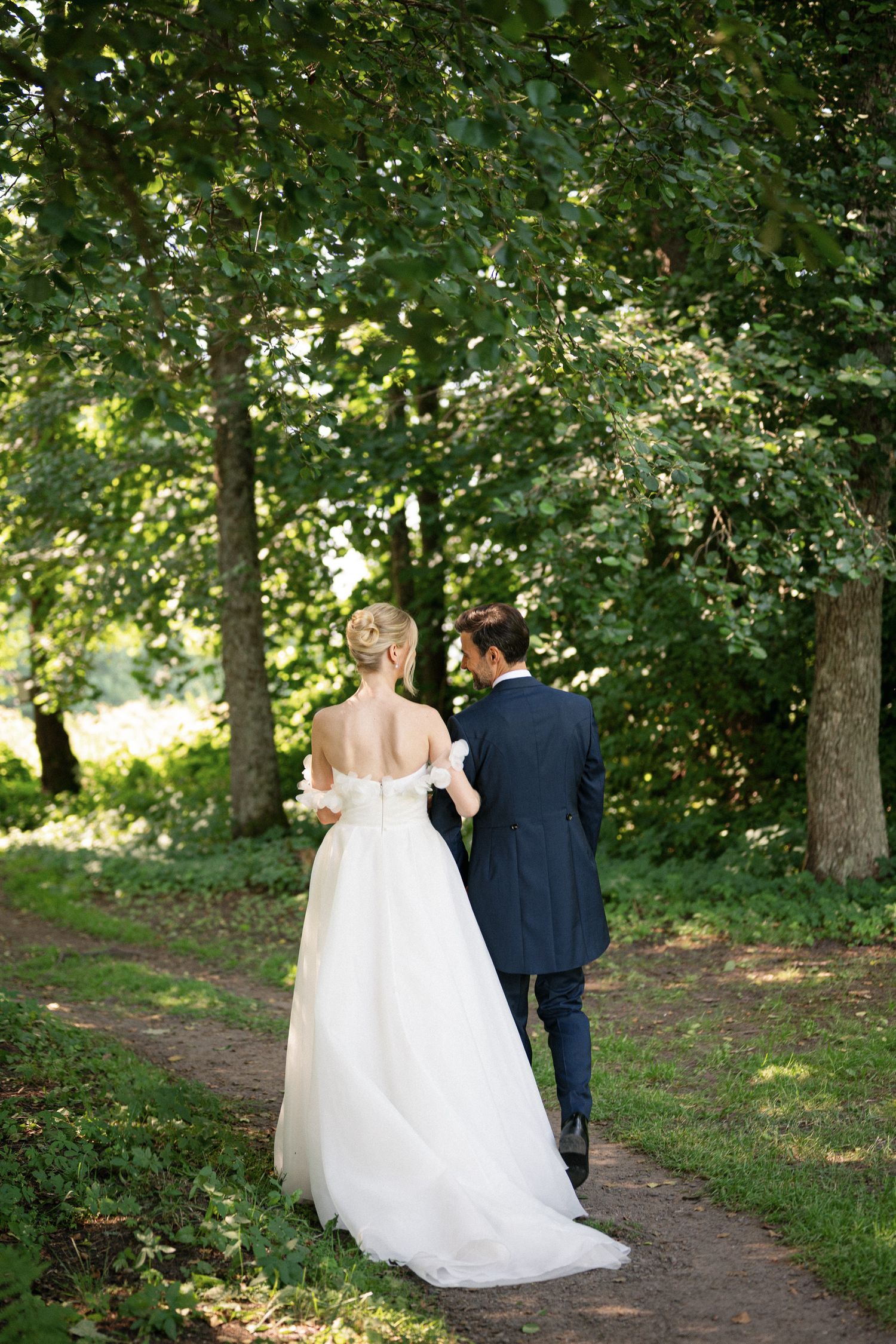 Bride in white backless gown and groom in navy suit walk hand in hand along a sunlit garden path surrounded by lush green trees.