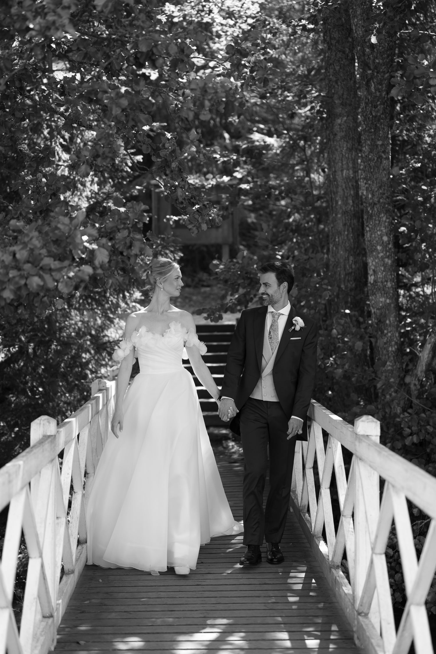 Black and white photo of a bride and groom holding hands walking on a wooden bridge surrounded by lush trees.