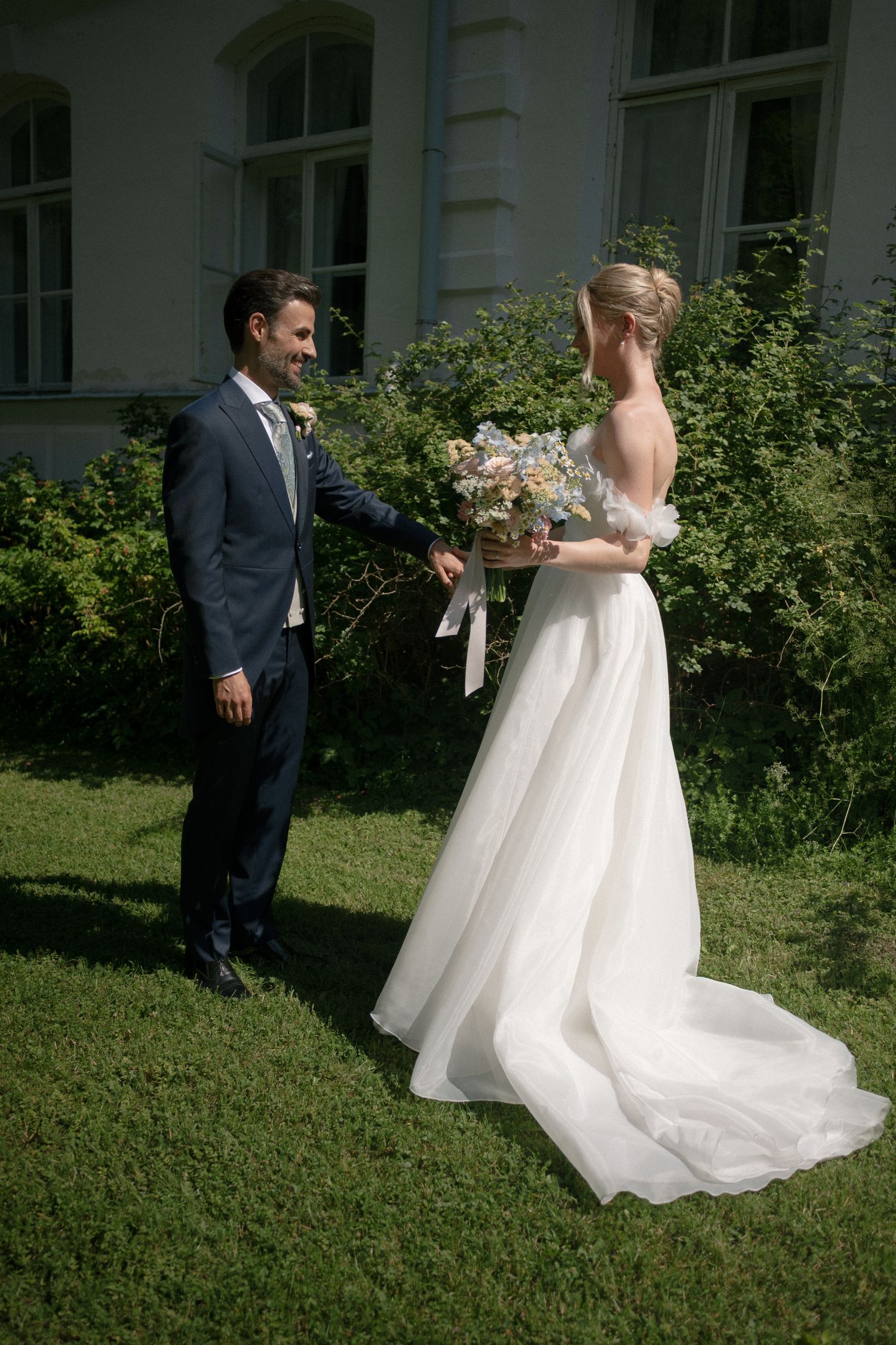 Bride in white gown holds bouquet as groom in navy suit reaches for her hand in a sunlit garden.