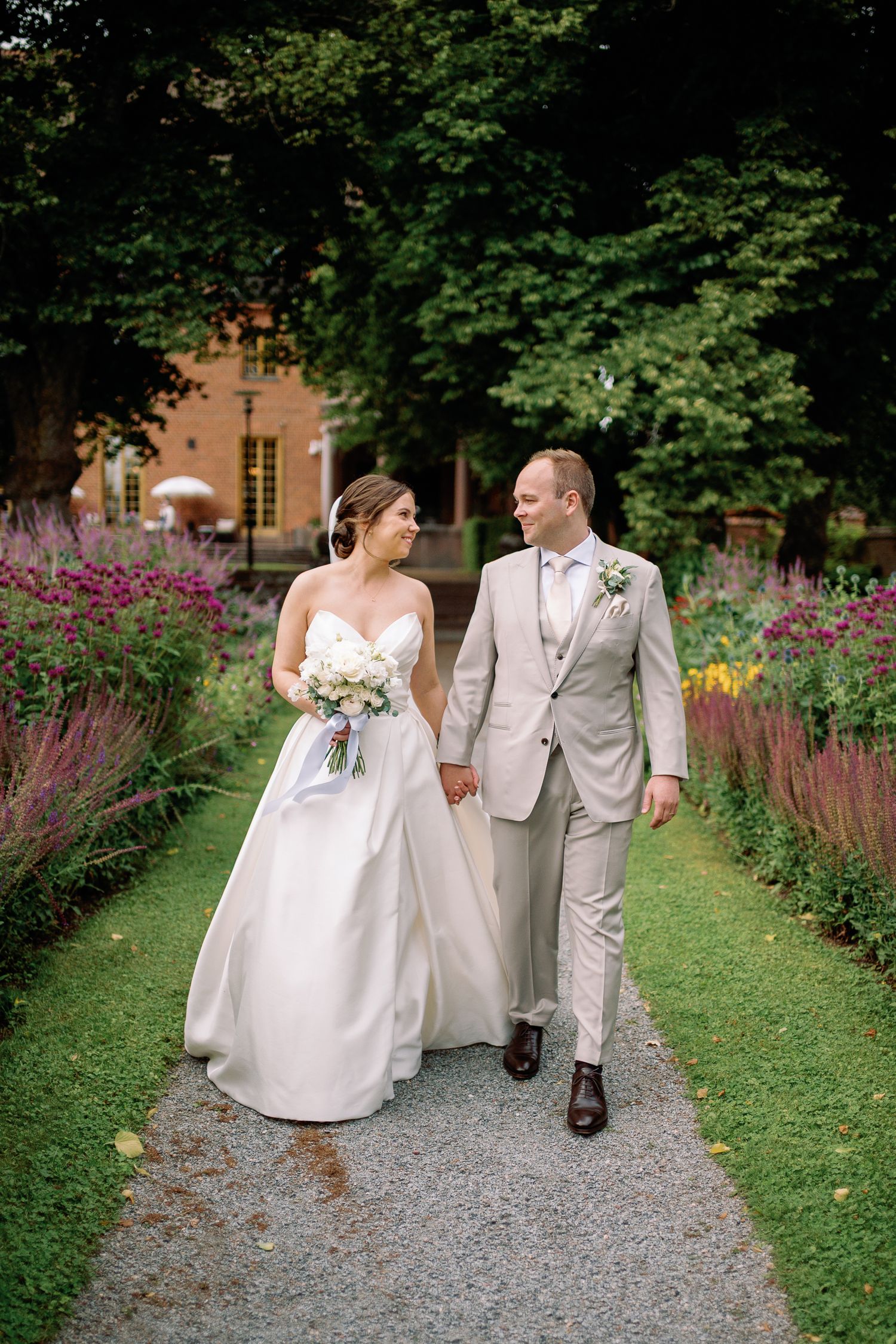 A bride and groom walk hand-in-hand along a garden path lined with colorful flowers on their wedding day.
