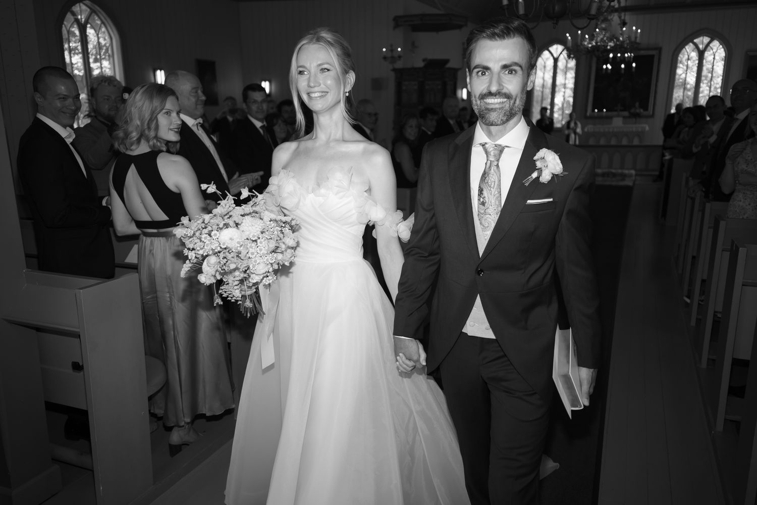 Black and white photo of a smiling bride holding bouquet and groom walking down church aisle after wedding ceremony.