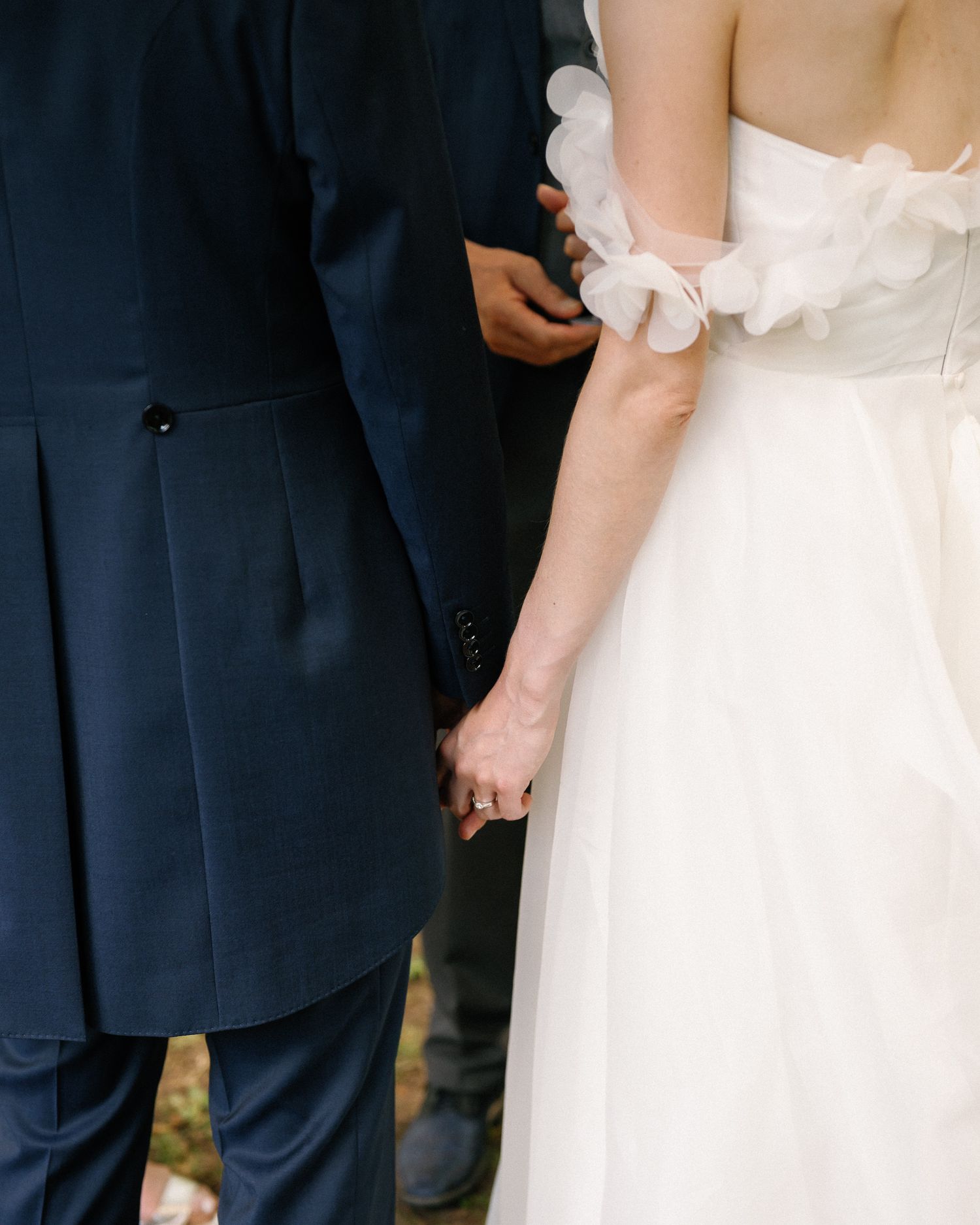 Bride and groom holding hands, wearing a white ruffled gown and navy suit, captured from behind at their wedding ceremony.