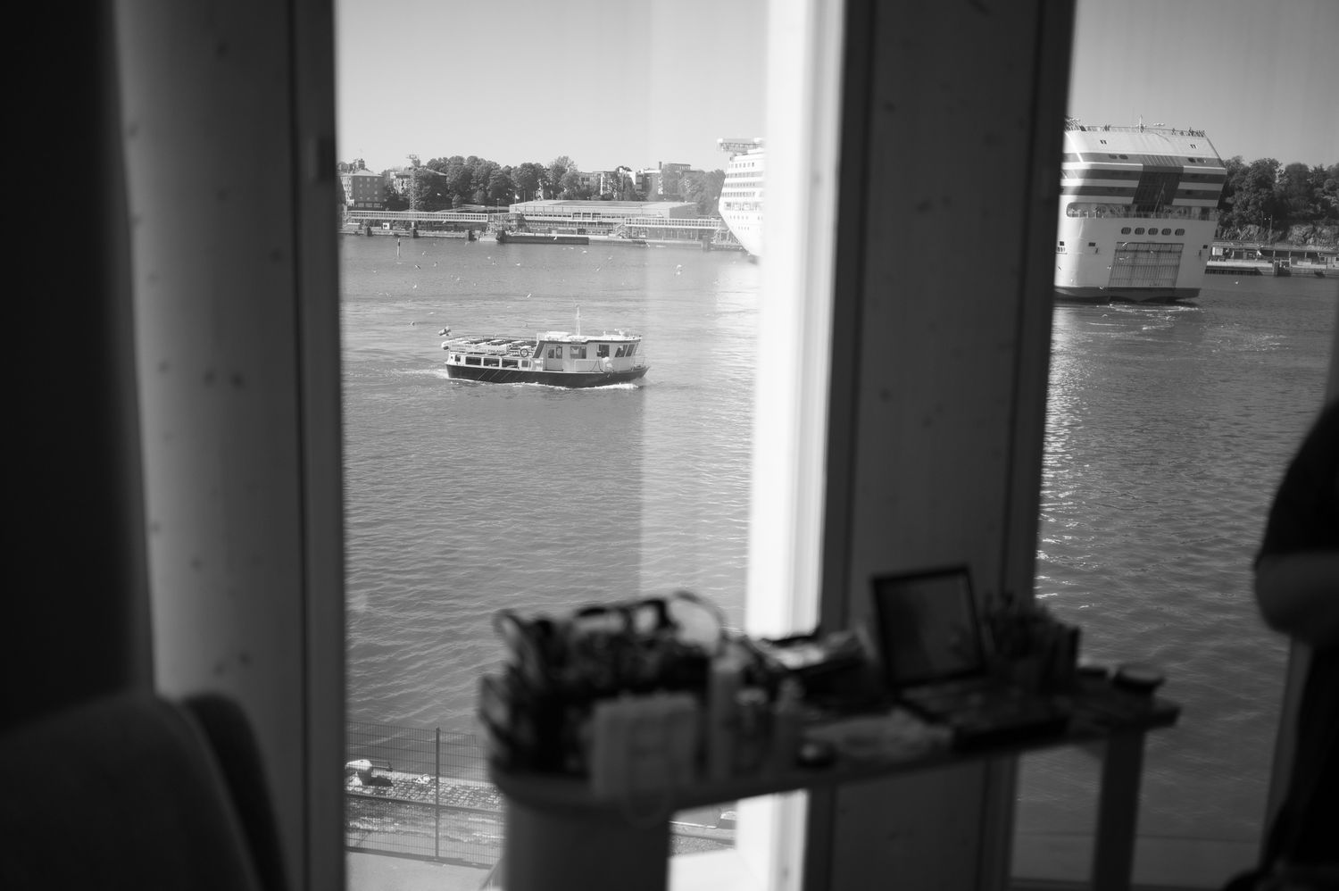 Black and white view through a window of a river with a ferry boat and large ships docked in the background.