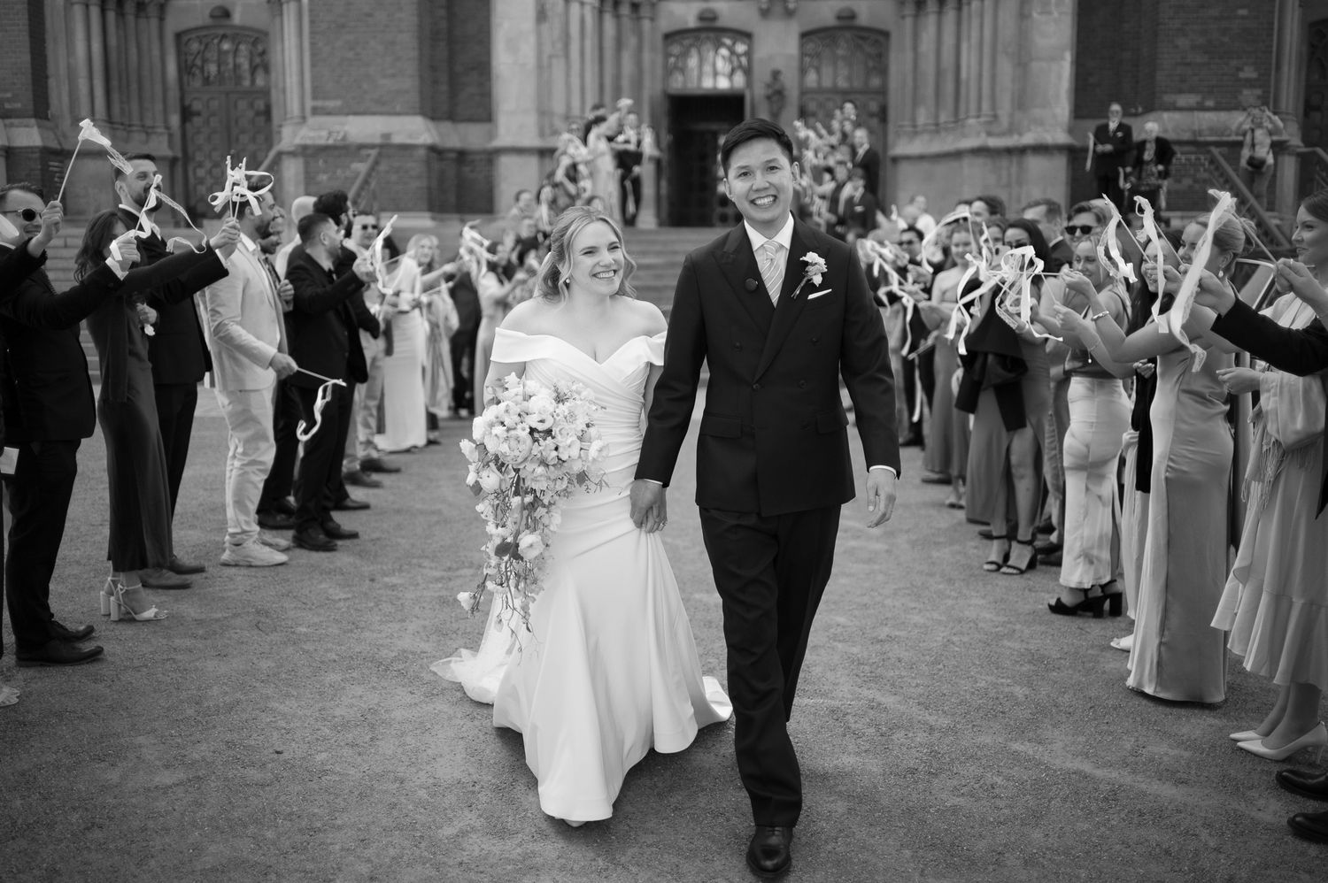 Black and white photo of a bride and groom walking through a celebratory arch of guests outside a church.