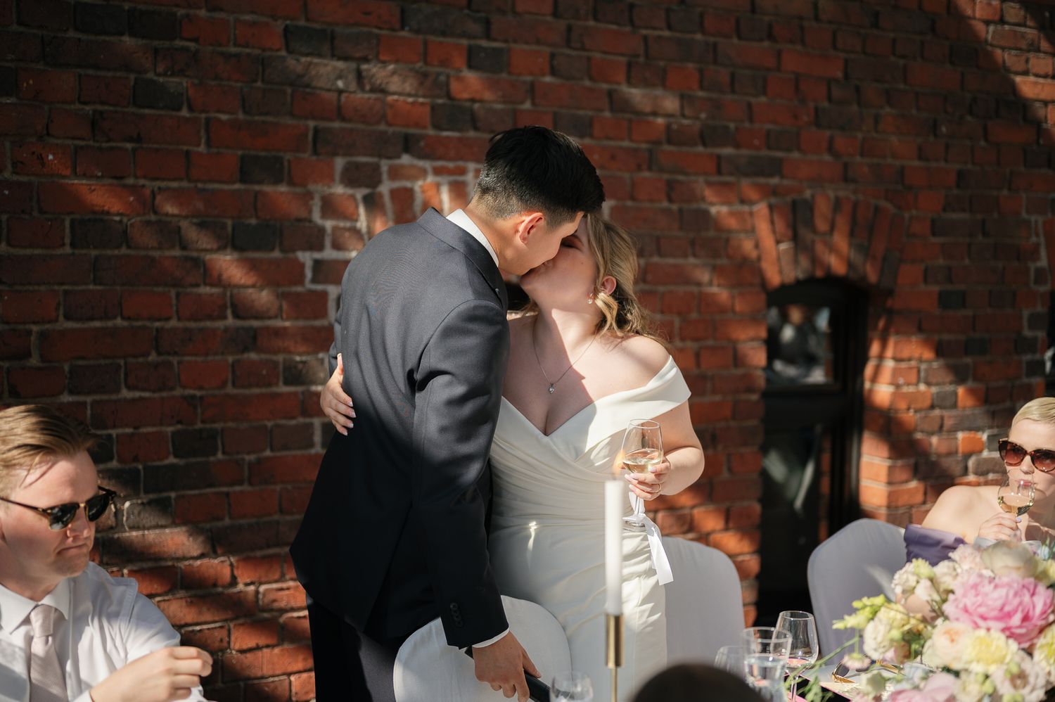 Bride and groom share a romantic first kiss at wedding reception with brick wall backdrop and floral arrangements.