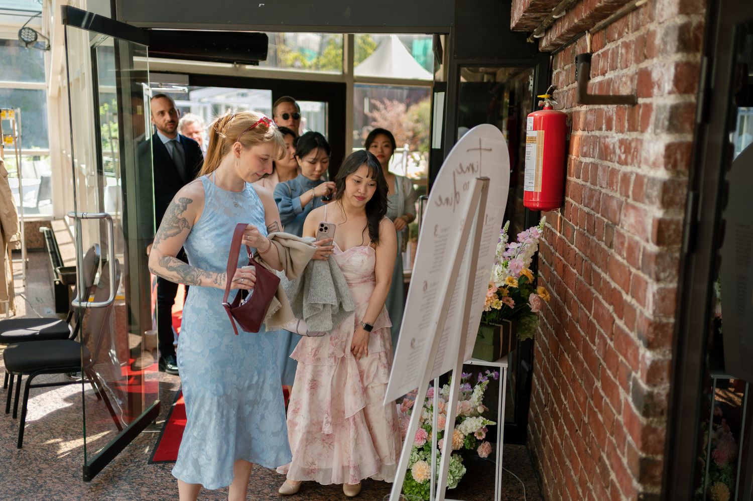 Guests in elegant dresses viewing a wedding sign board at a venue entrance with brick walls and floral decorations.