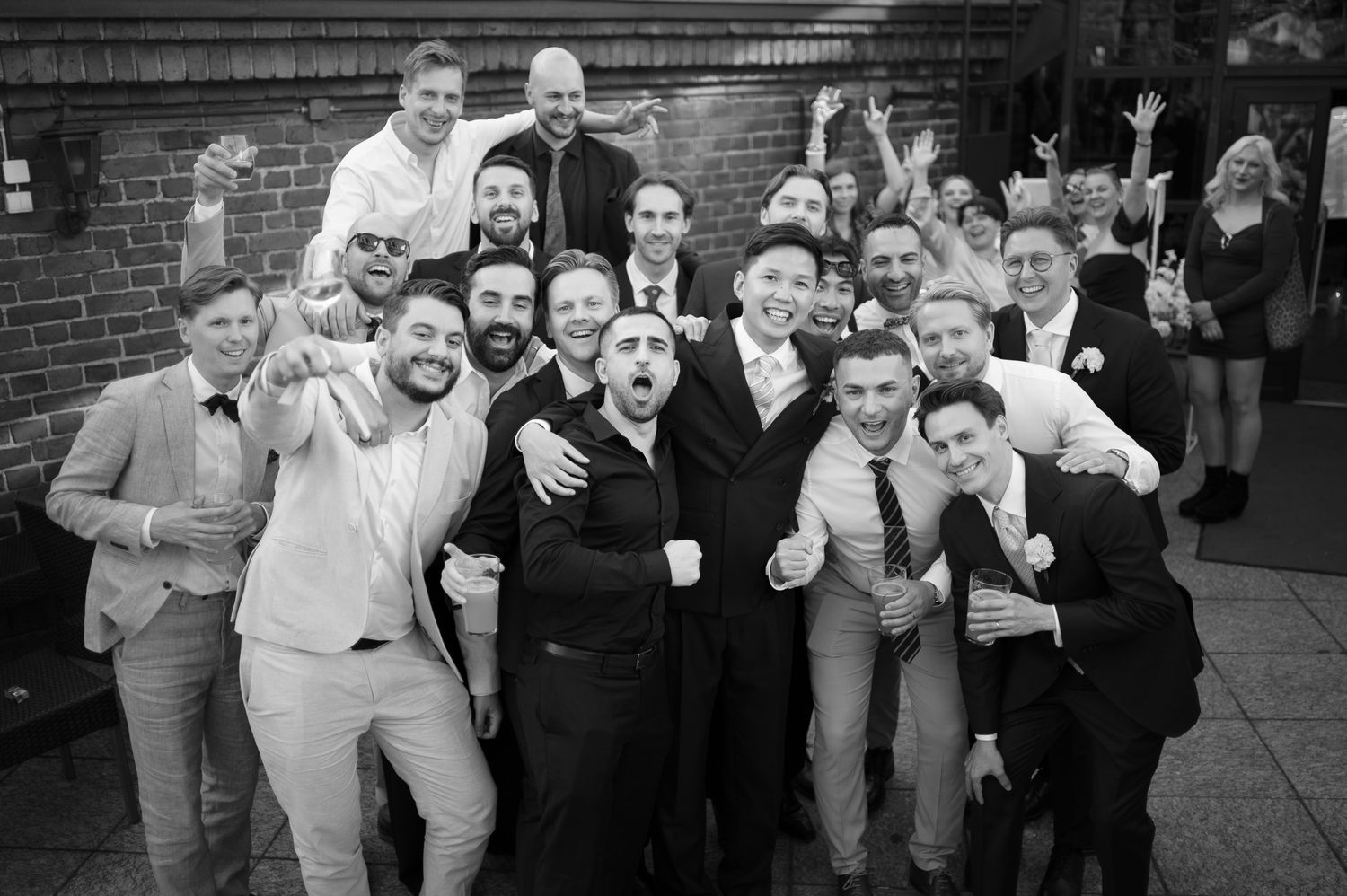 Black and white photo of a large group of men in suits celebrating together at a wedding reception outdoors.