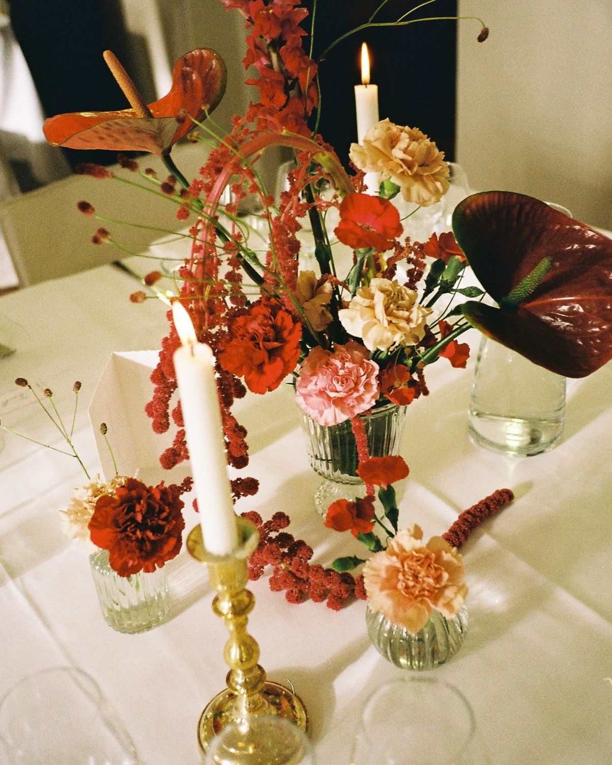 Elegant floral centerpiece with red, pink carnations, candles, and tropical blooms in glass vases on a white tablecloth.