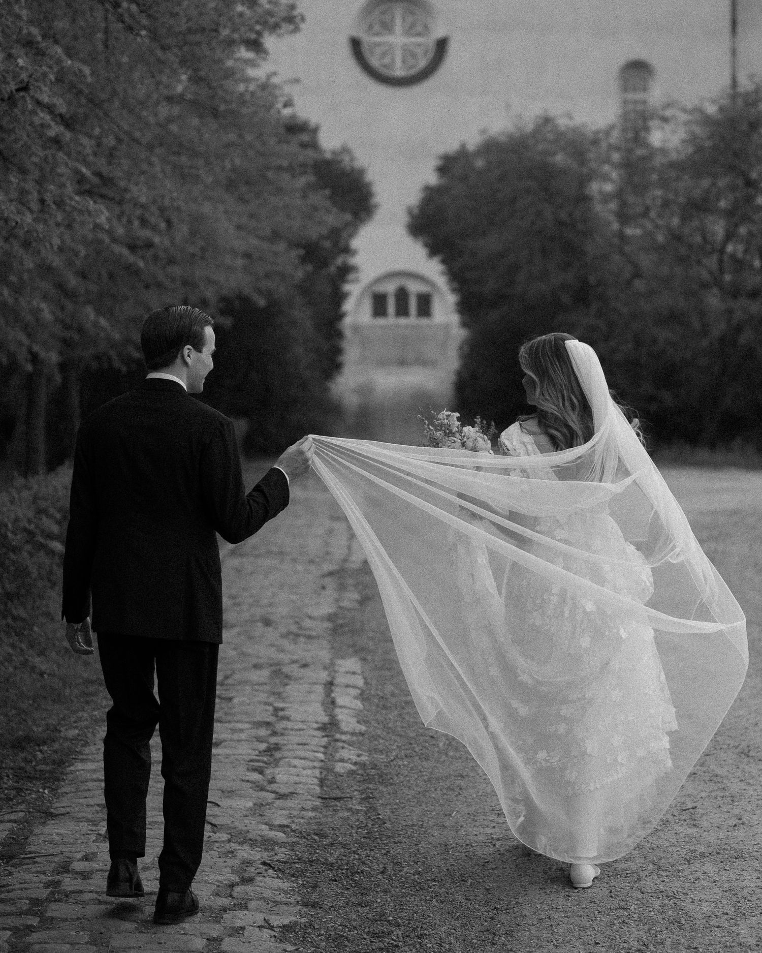 Black and white photo of a bride and groom walking away, her veil flowing in the wind toward a church.