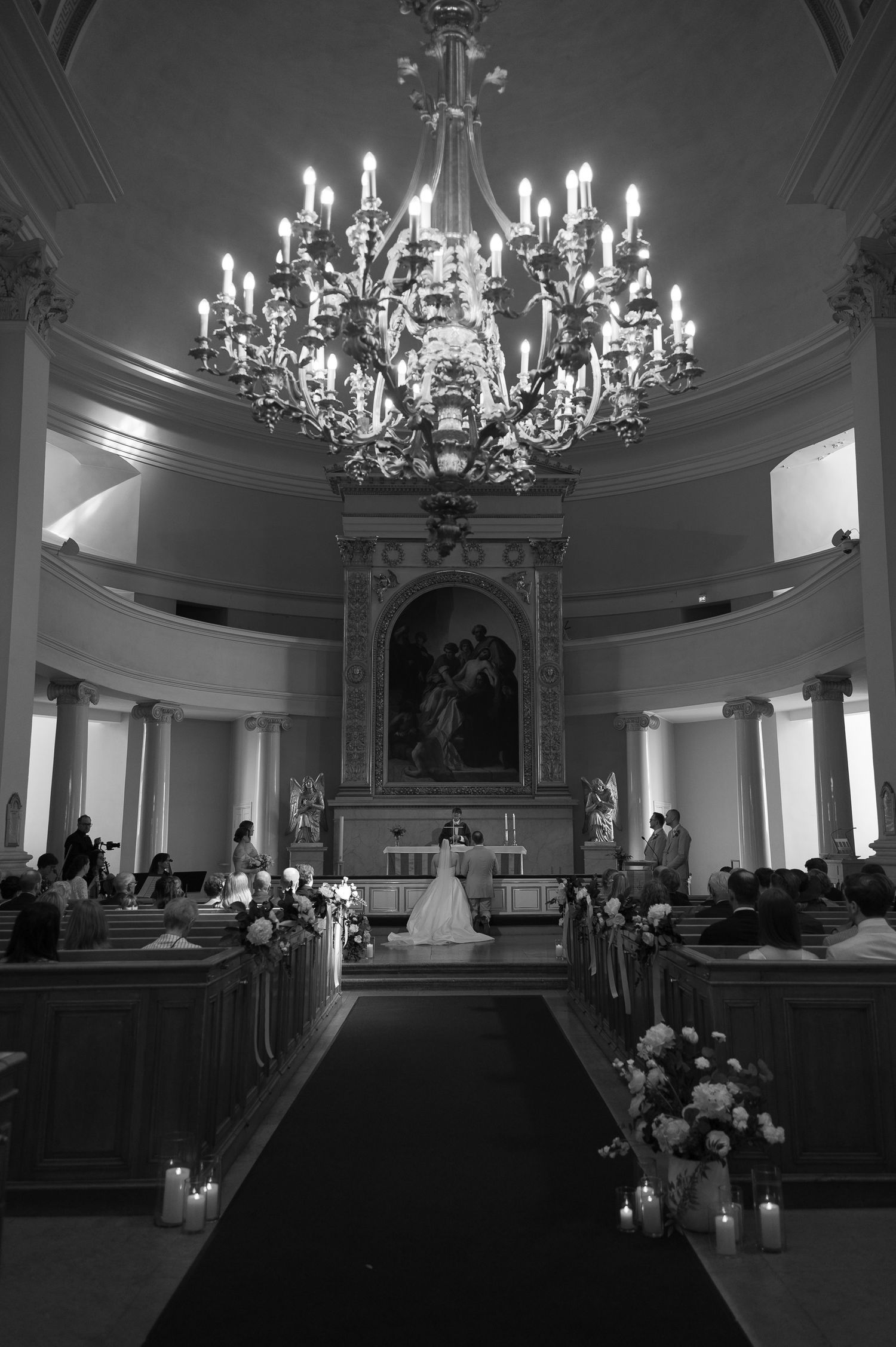 Black and white photo of an elegant church wedding ceremony with chandelier, altar, and floral arrangements lining the aisle.