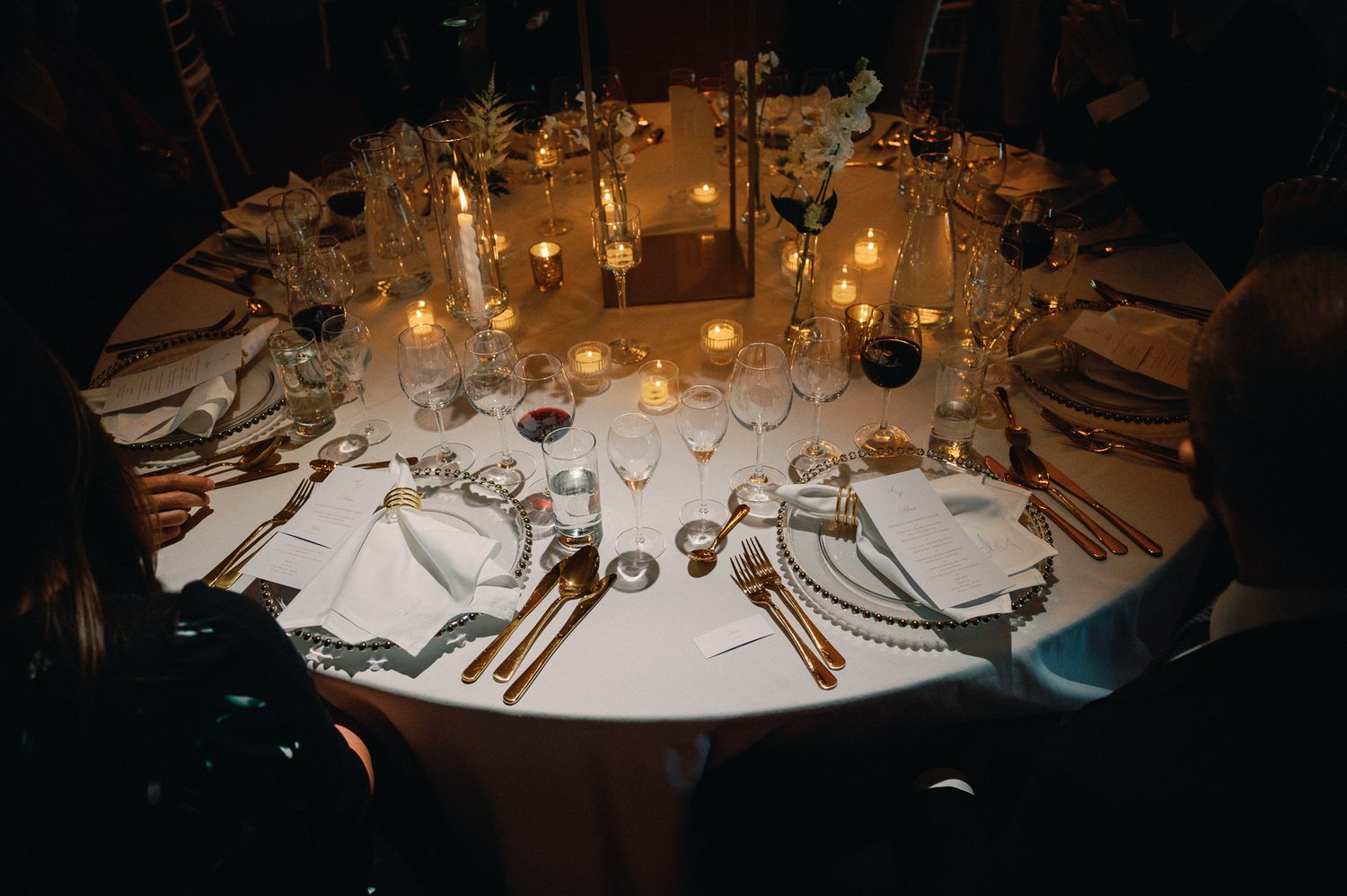 Elegant round wedding reception table set with candles, fine china, and golden cutlery in a dimly lit venue.