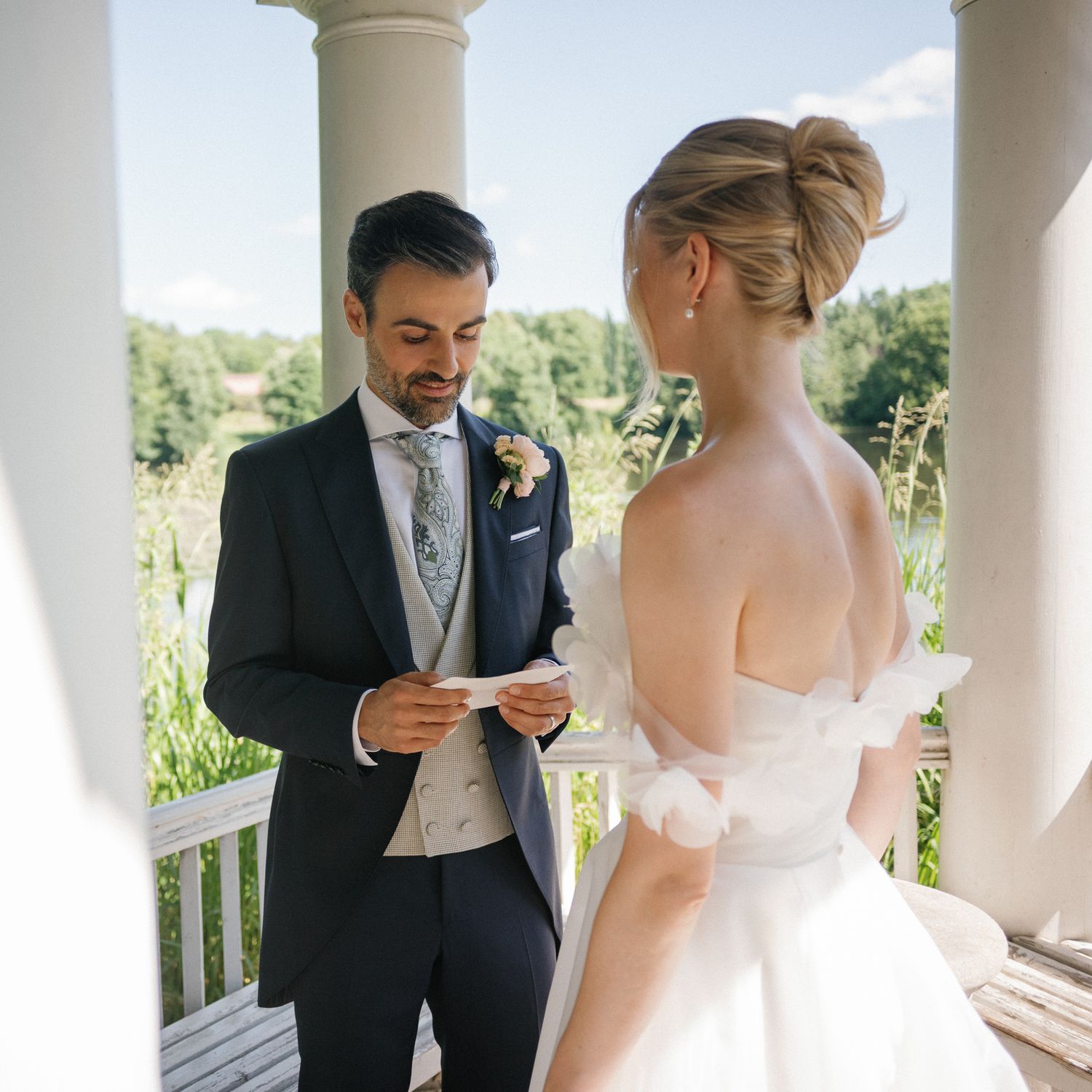 Groom reads wedding vows to bride in elegant off-shoulder dress at outdoor ceremony with white columns.