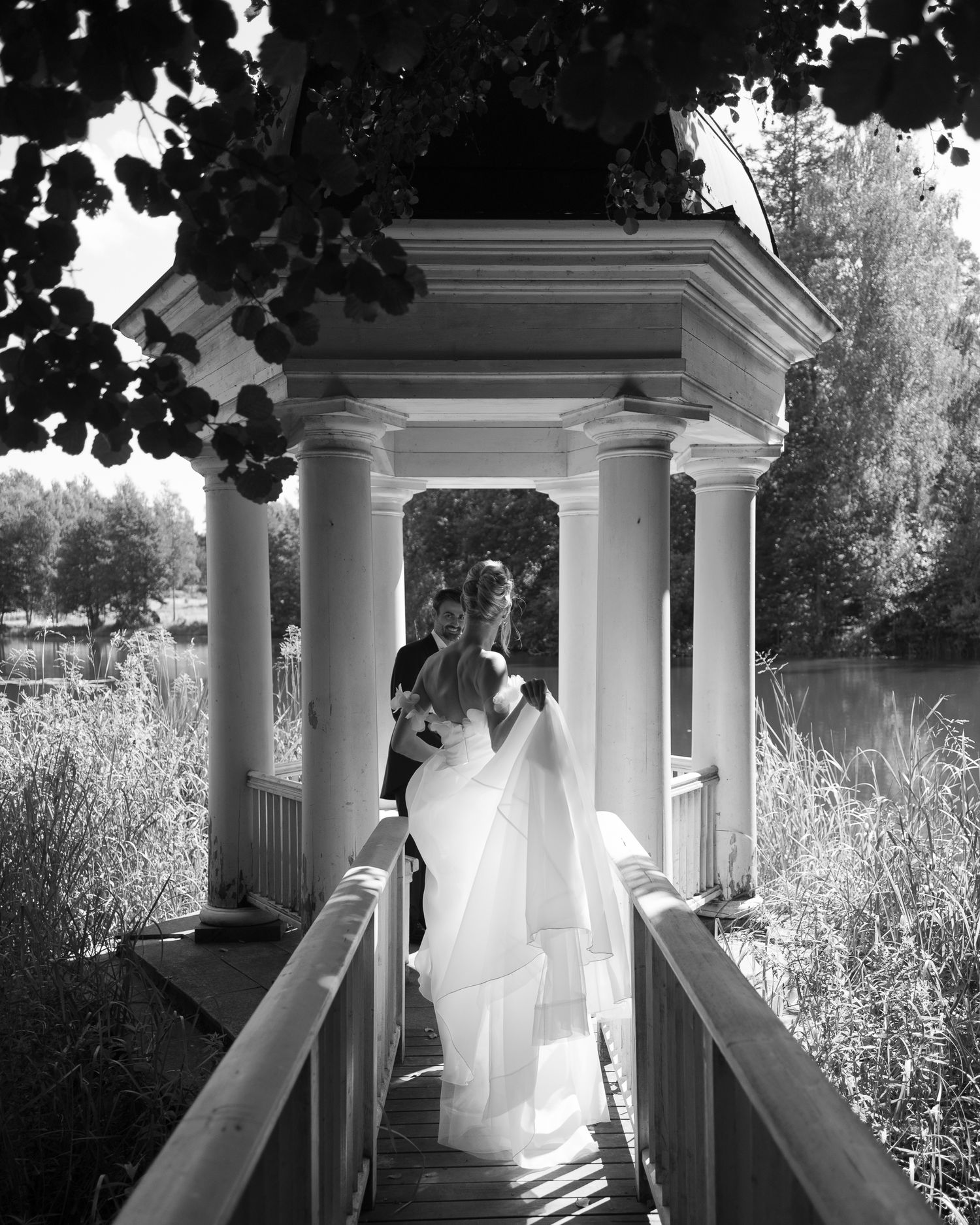 Black and white photo of a bride and groom standing under a classical gazebo by a lake on their wedding day.