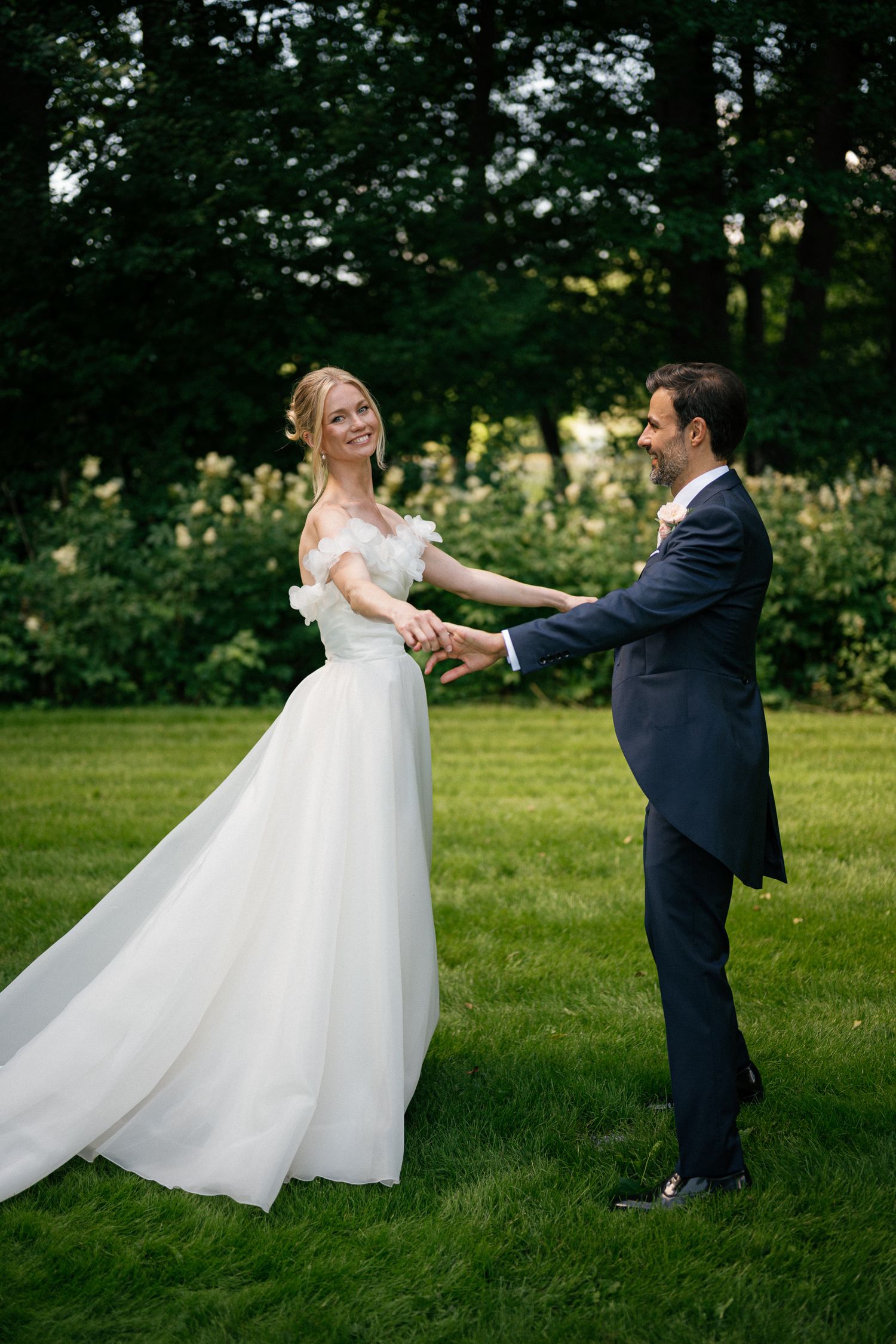 A bride in a white off-shoulder gown holds hands with a groom in a navy suit on a lush green lawn.