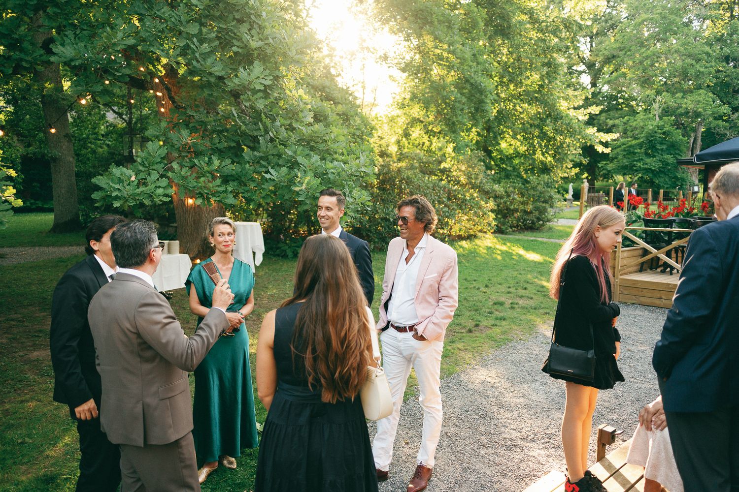 Guests mingle outdoors at a garden party during golden hour, surrounded by lush green trees and warm sunlight.
