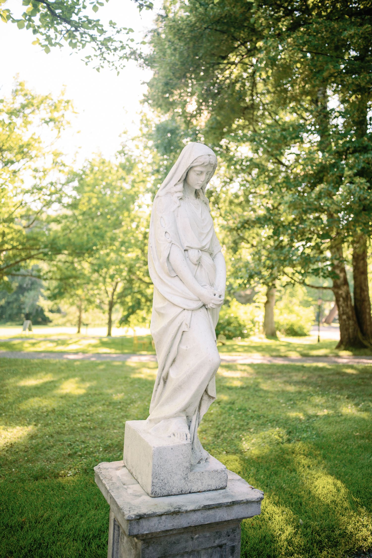 White marble Virgin Mary statue on stone pedestal in sunlit green park with tall trees.