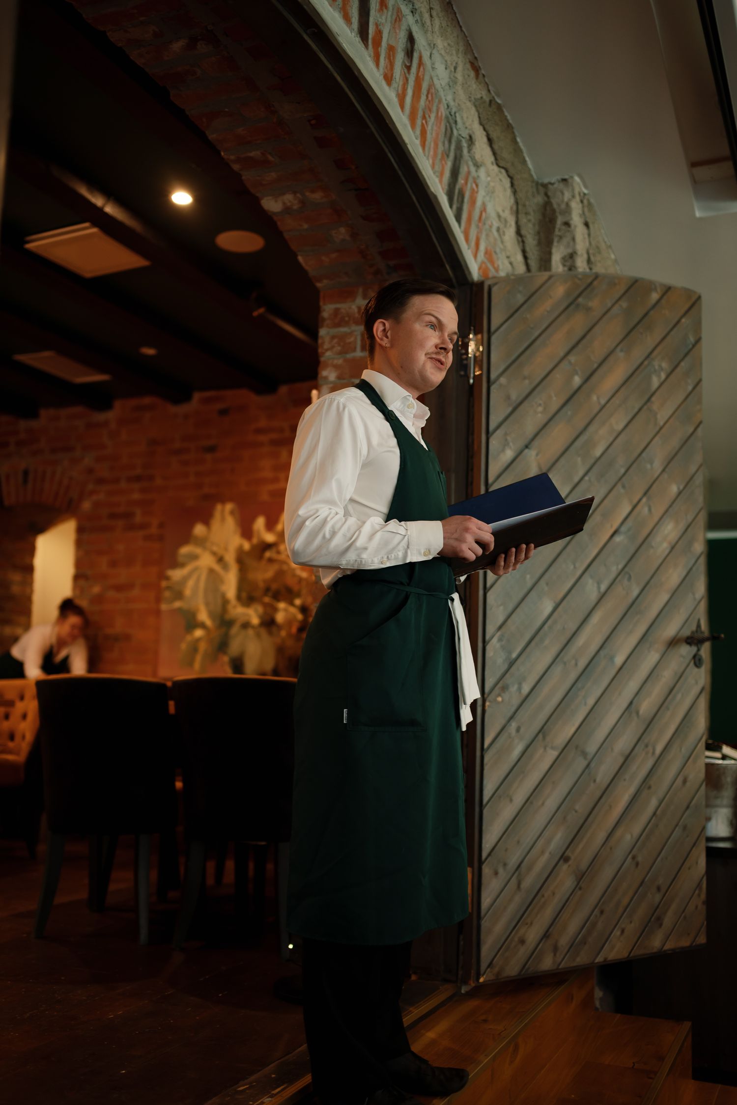 A waiter in a green apron holds a menu while standing near a wooden door in an elegant restaurant.