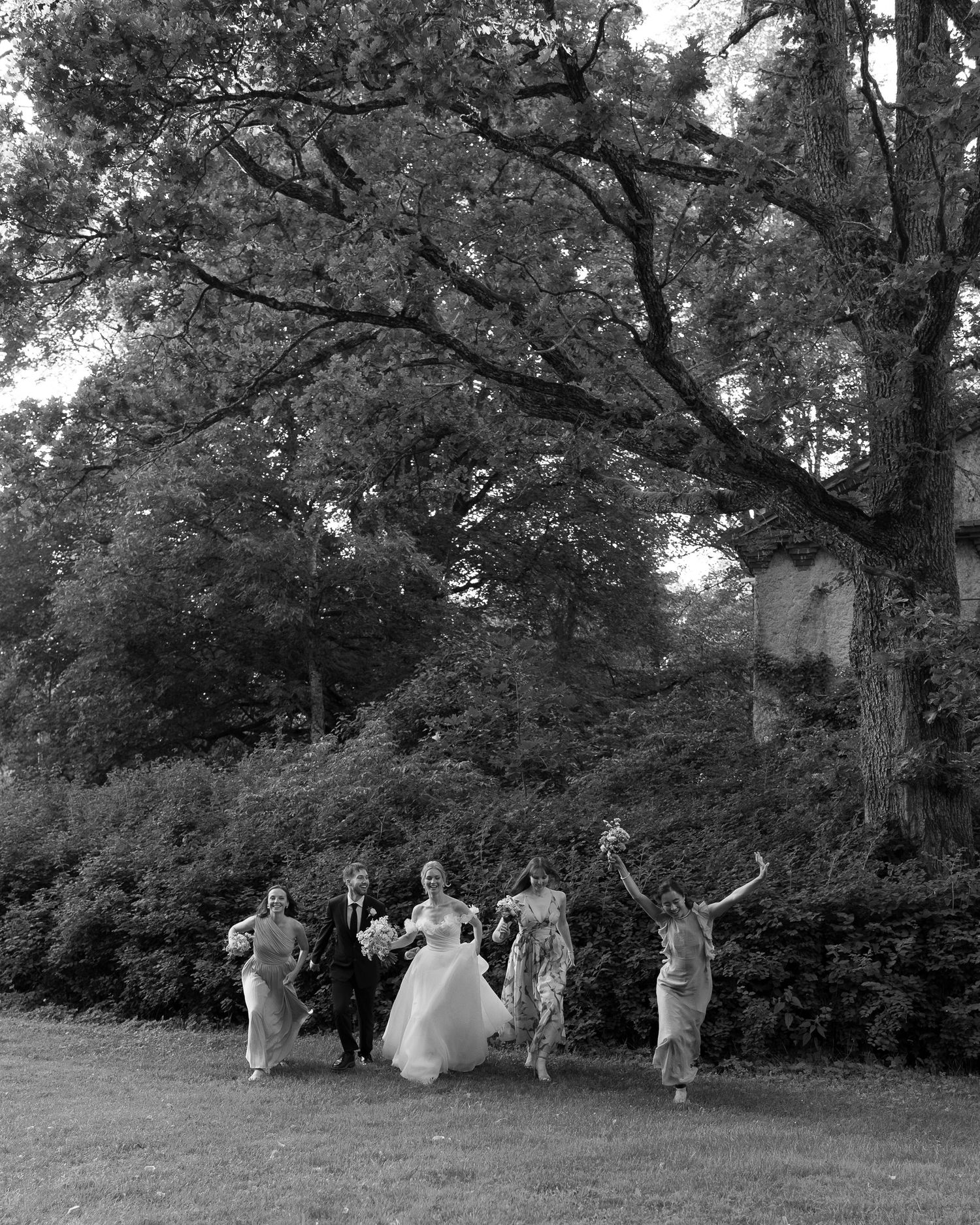 Black and white photo of a bride, groom, and bridesmaids joyfully jumping outdoors under large trees.
