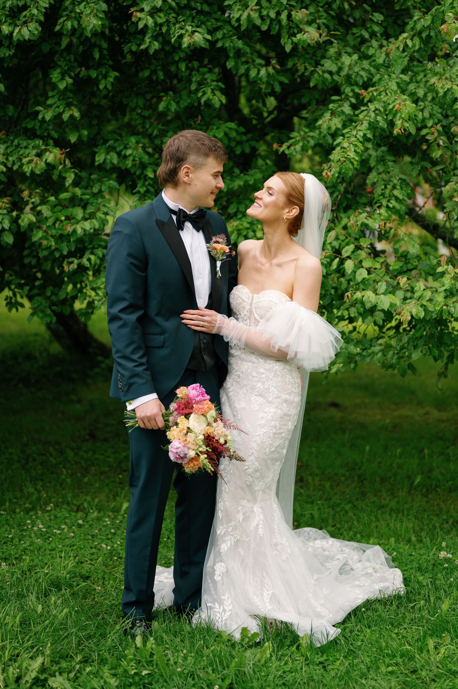 Romantic wedding couple embracing on grass with lush green trees in the background.