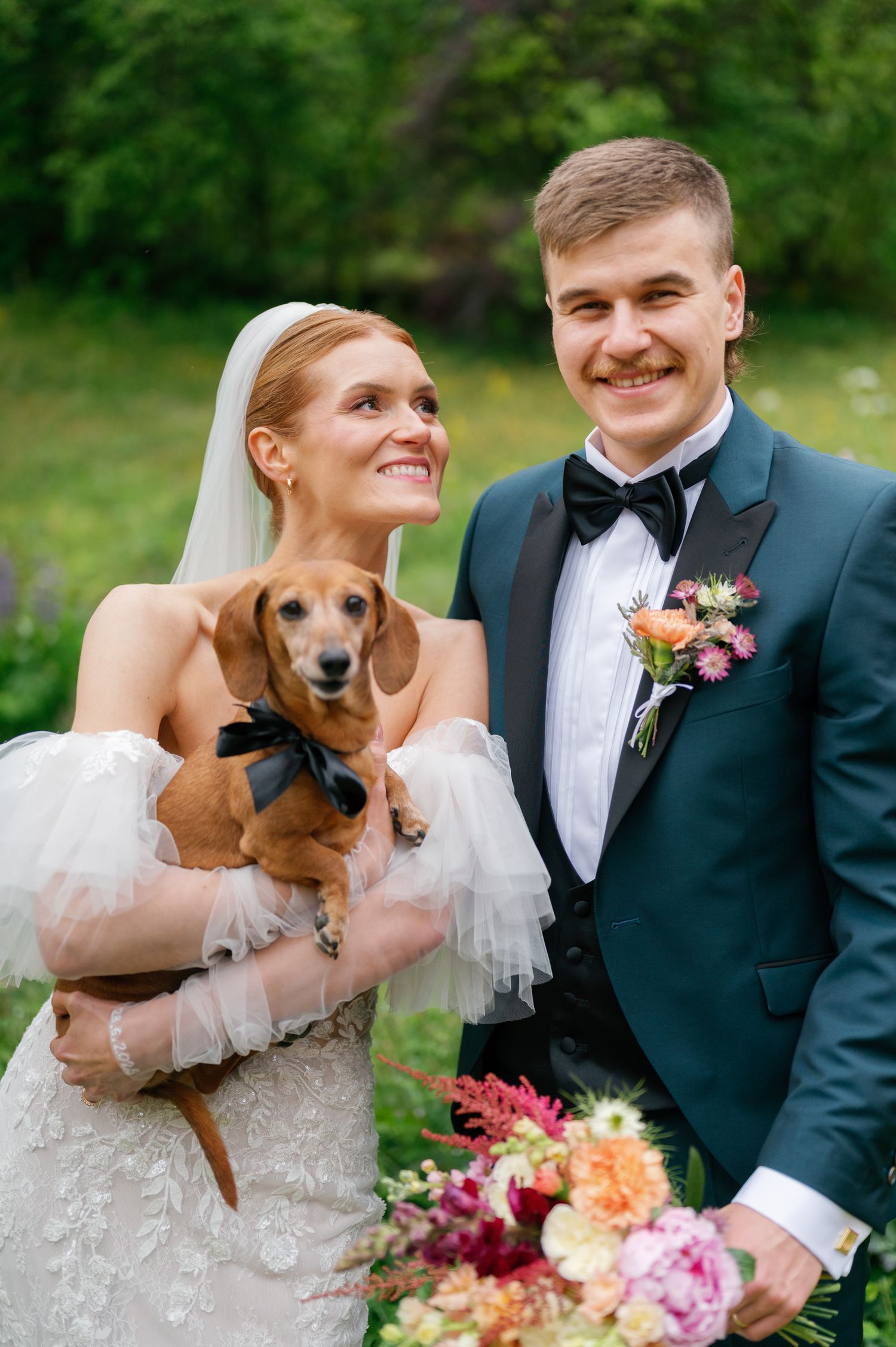 A couple in wedding attire holds a small brown dachshund dog during their outdoor photo session.