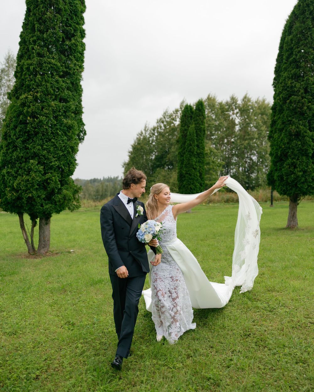 Bride's veil flows in the wind as couple walks through garden setting.