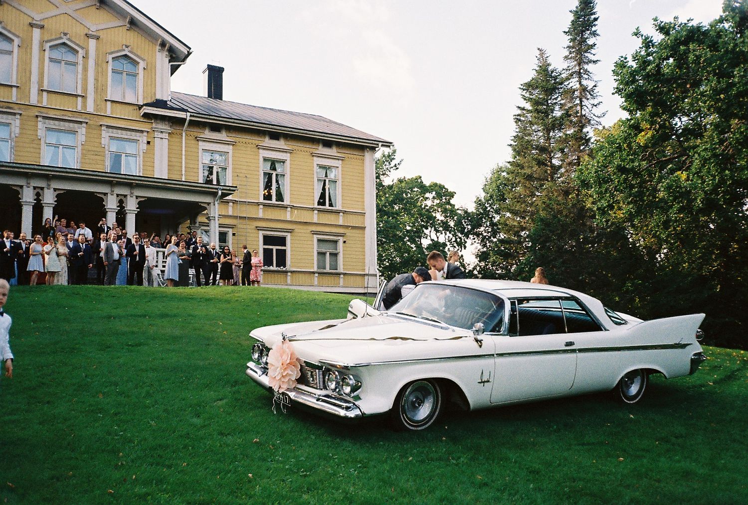 Classic white vintage car parked on lawn in front of elegant yellow mansion.