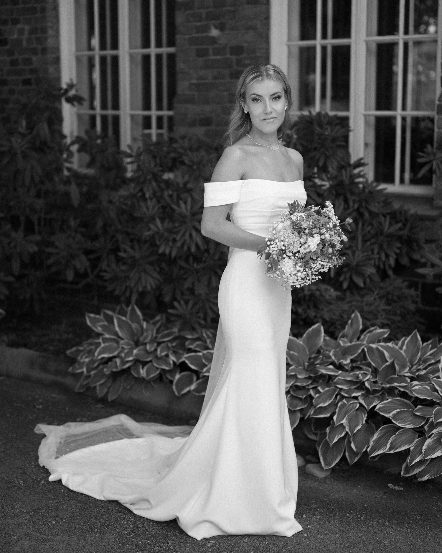 A bride in an elegant off-shoulder white wedding gown stands in front of a brick building with lush greenery.