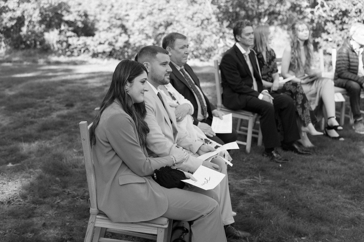 Wedding guests seated on chairs on a grassy lawn during an outdoor ceremony.