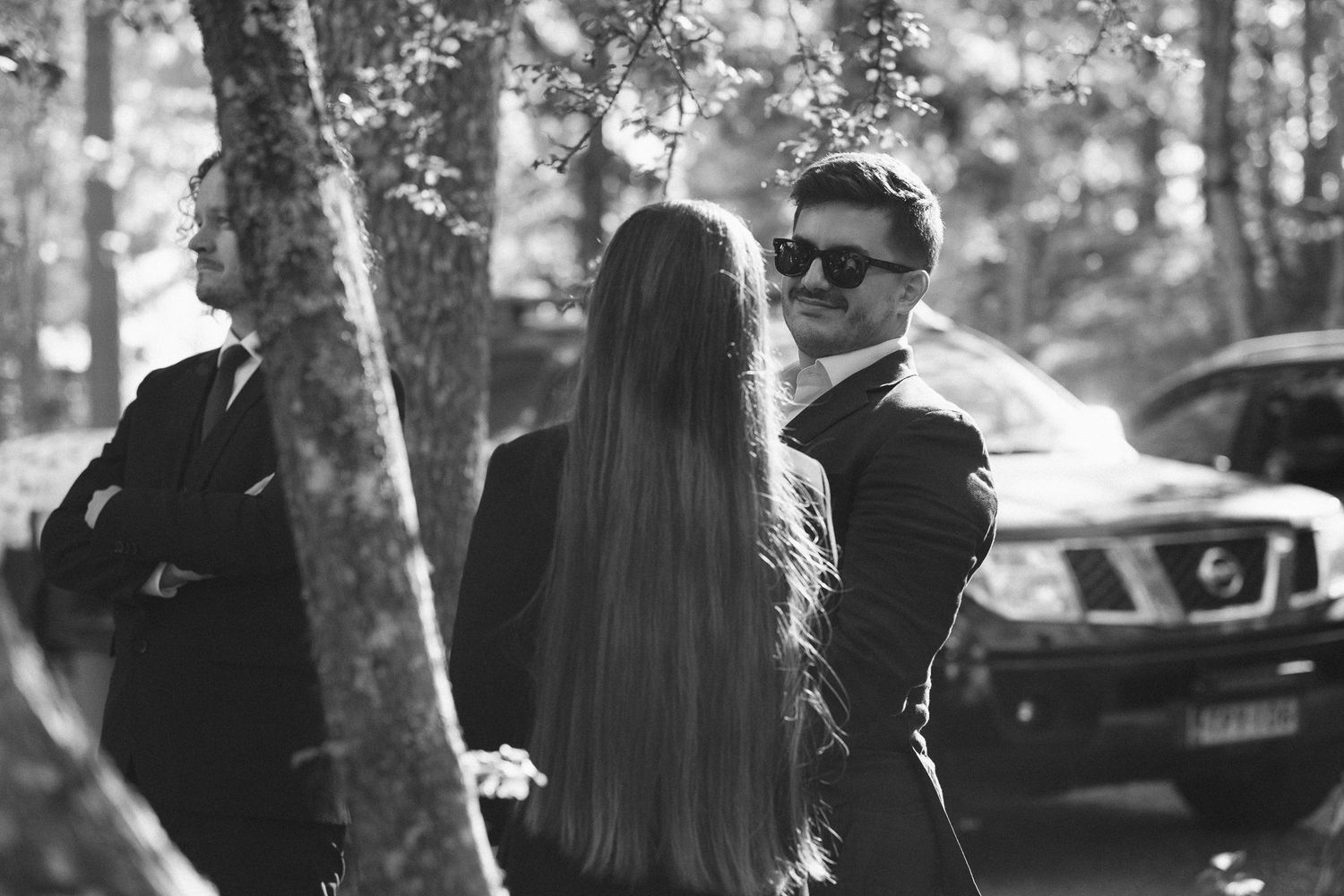 Wedding guests mingle outdoors among trees in formal attire.
