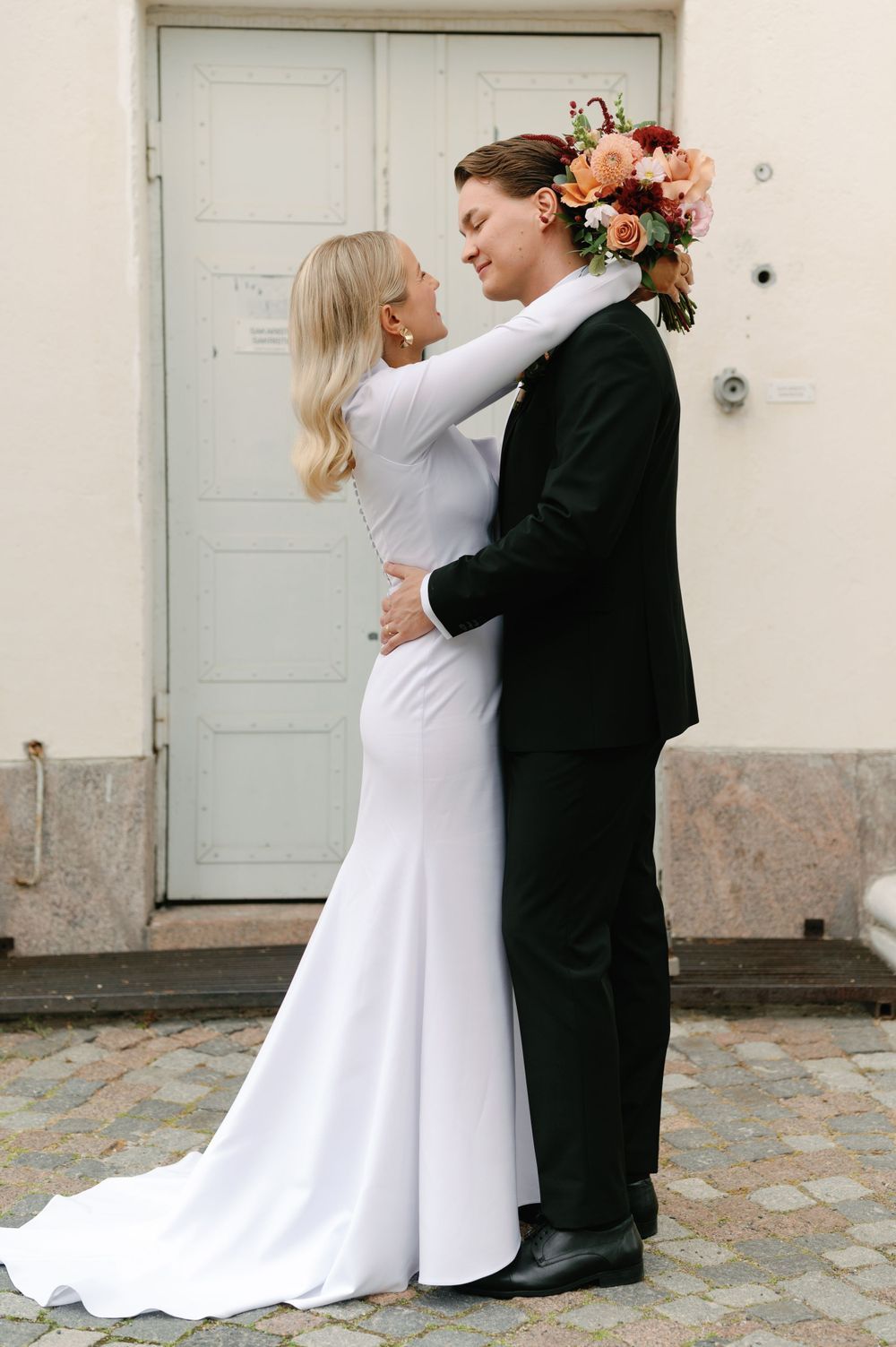 A couple in wedding attire share an intimate moment in front of a white door with a colorful floral bouquet.