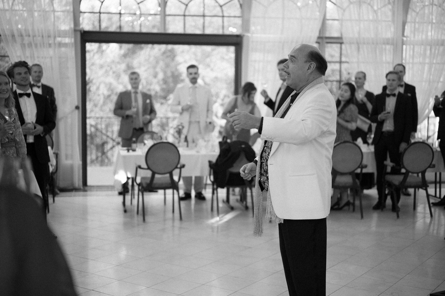 Black and white photo of wedding guests gathered in an elegant venue with floor-to-ceiling windows.