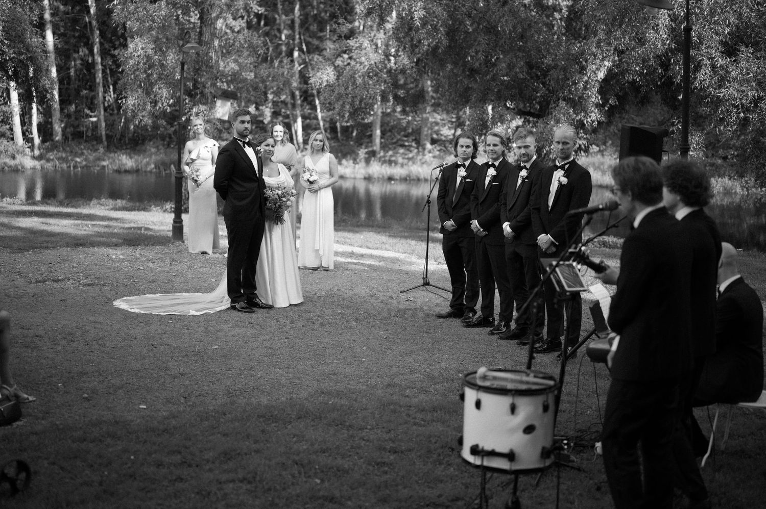 Wedding party members gather on a gravel path surrounded by lush greenery at an outdoor venue.