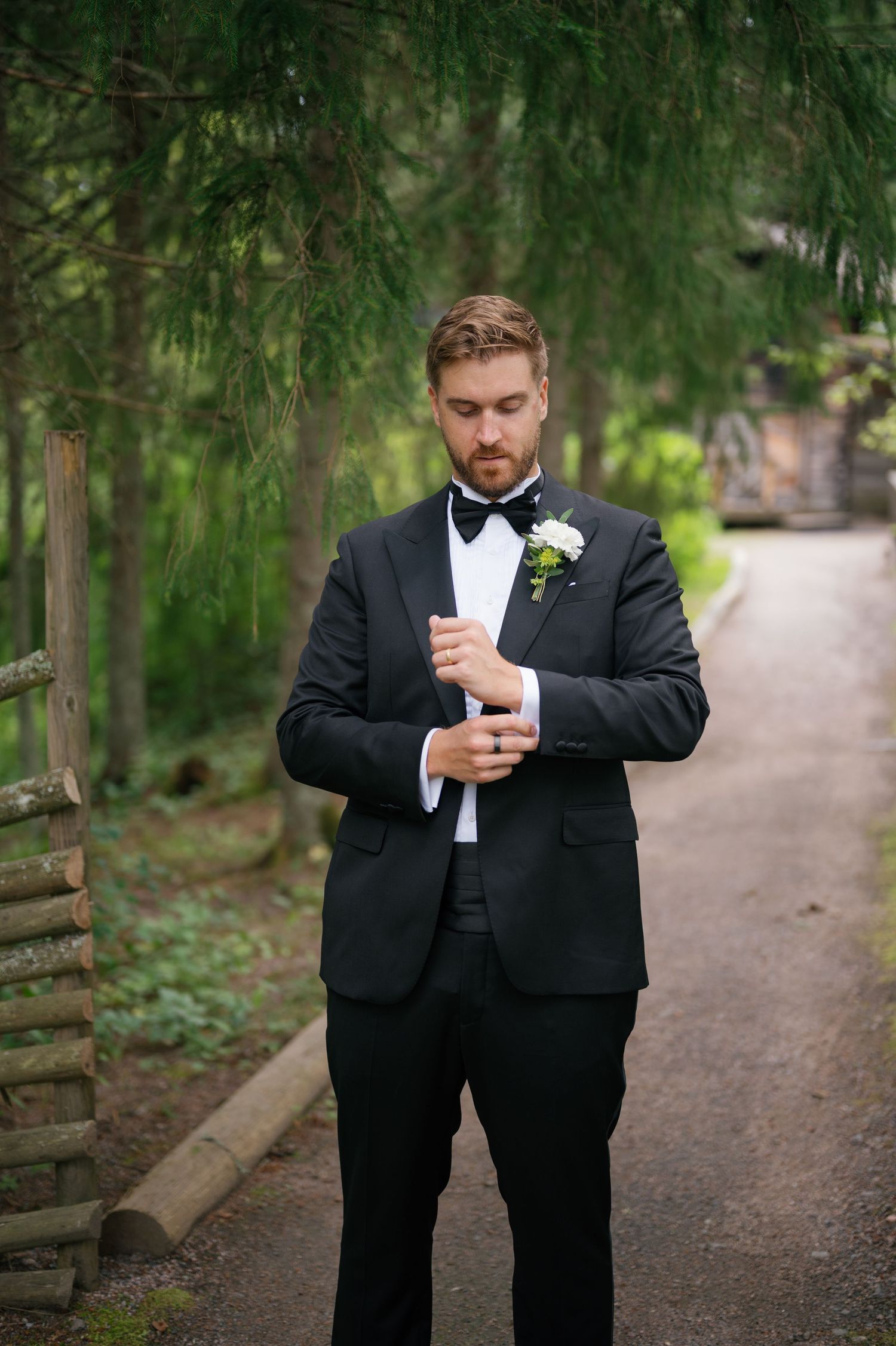 Groomsmen in formal black tuxedos checking their phones on a wooded path.
