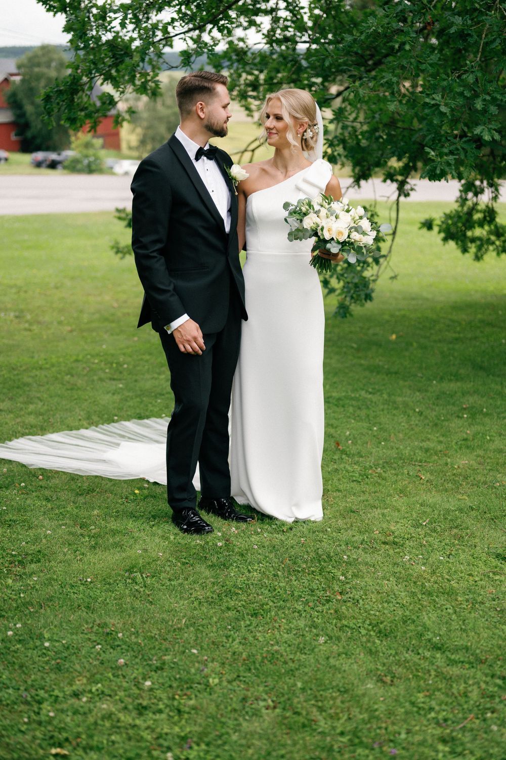 A wedding couple walks together on lush green grass beneath overhanging tree branches.