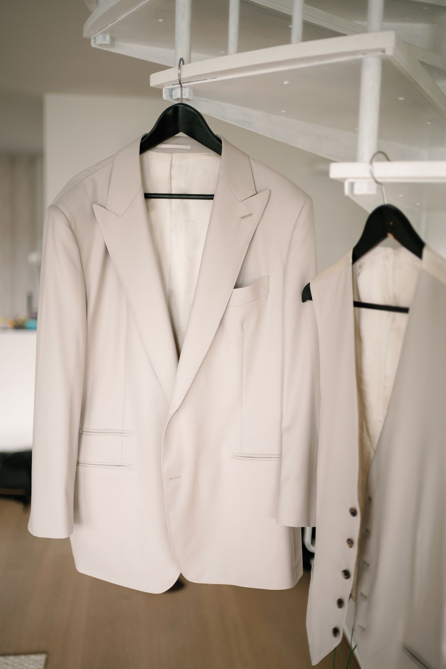 White wedding suit jacket hanging on a black hanger, with a wedding dress visible in the background.