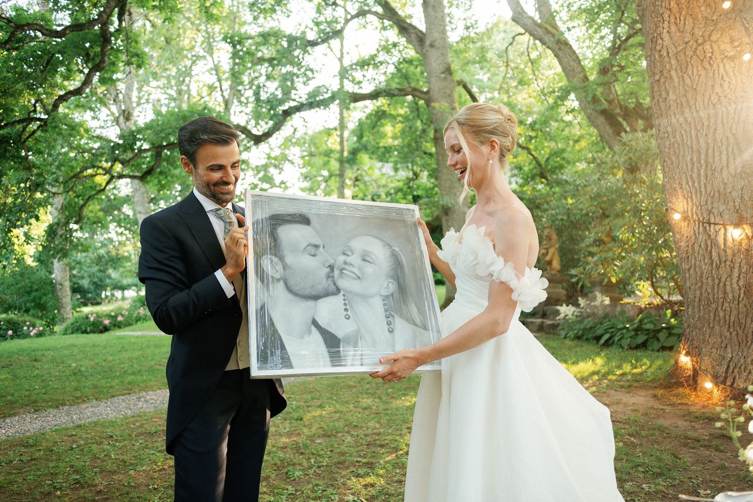 Bride and groom holding a framed black and white portrait sketch at outdoor wedding ceremony surrounded by lush green trees.