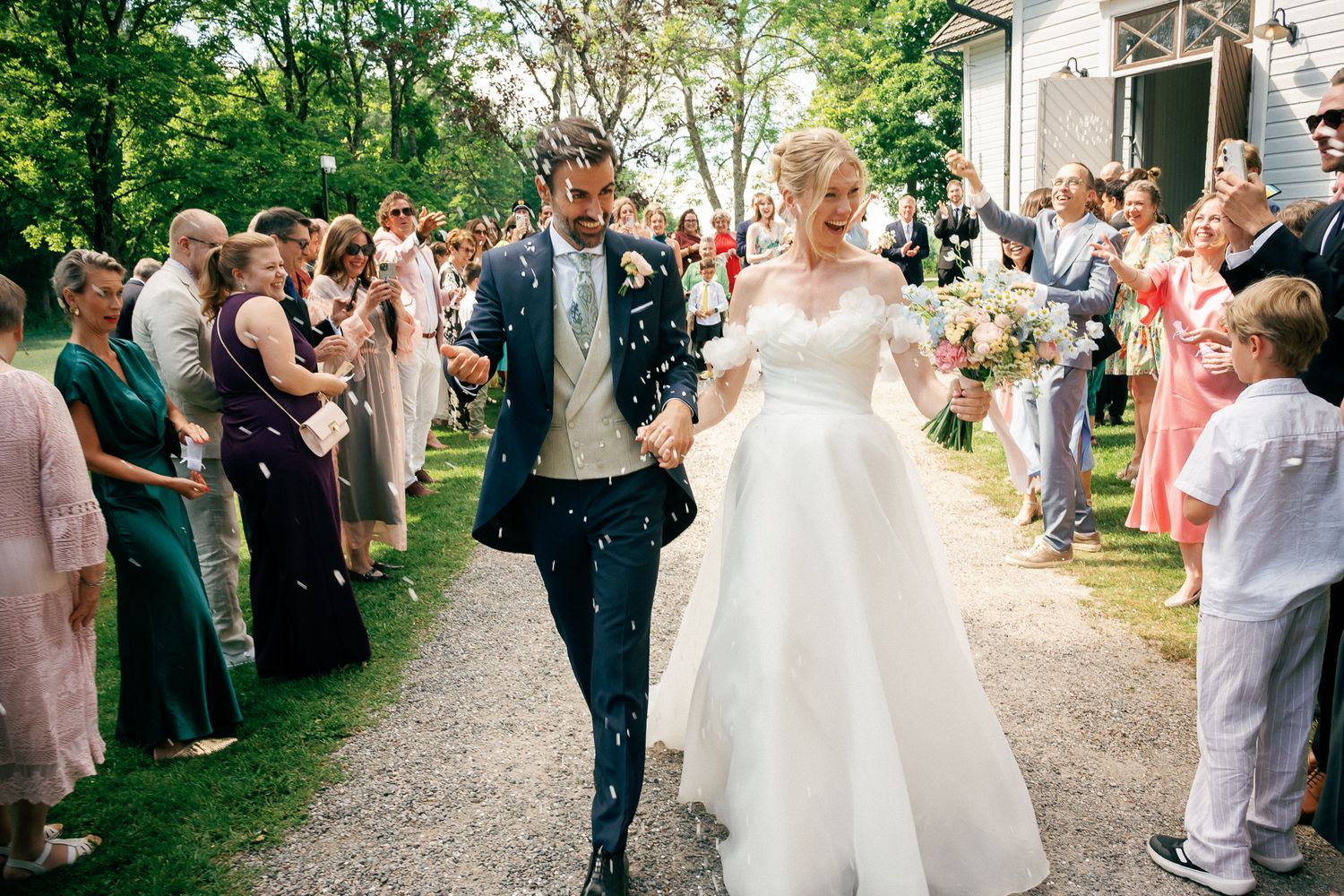 Bride and groom walk through confetti shower surrounded by cheering wedding guests on a tree-lined path.