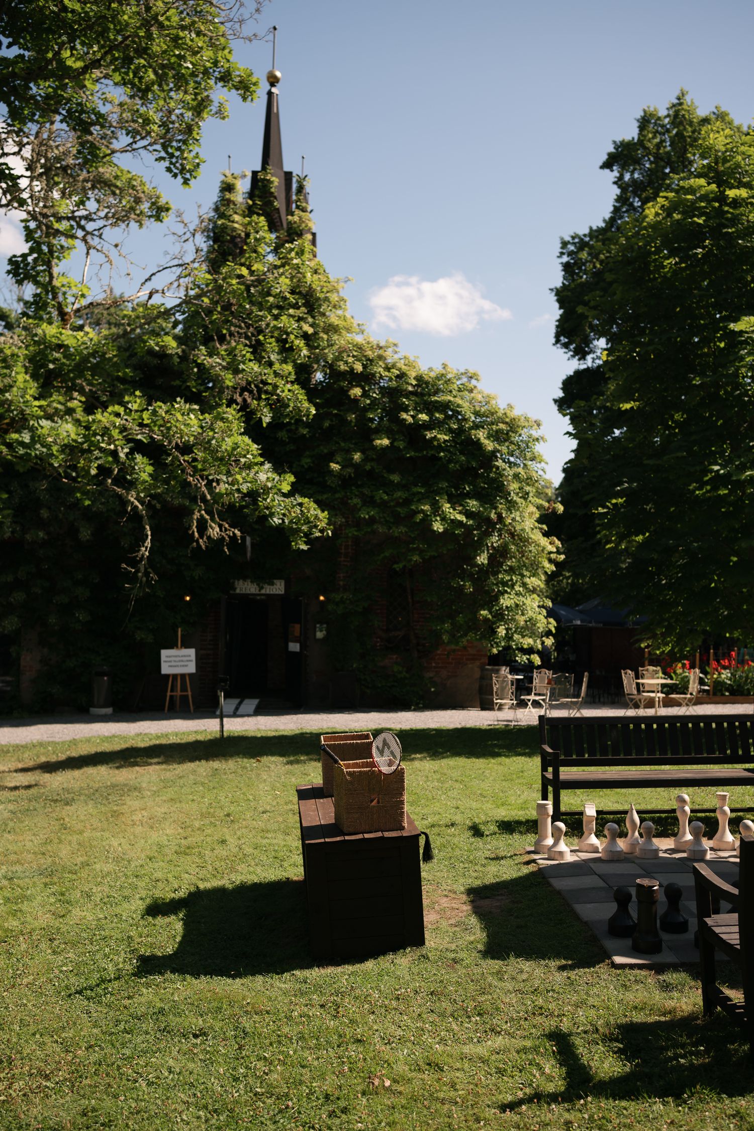 Outdoor garden with giant chess set on grass, brick church with steeple, and lush green trees under a blue sky.