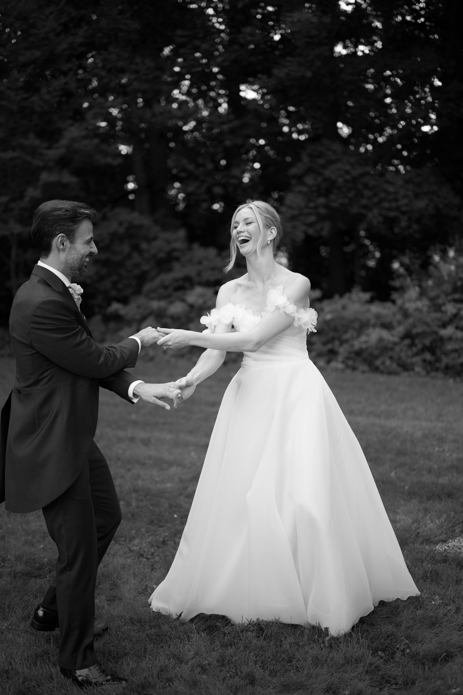Black and white photo of a laughing bride in off-shoulder gown dancing with groom in suit outdoors.