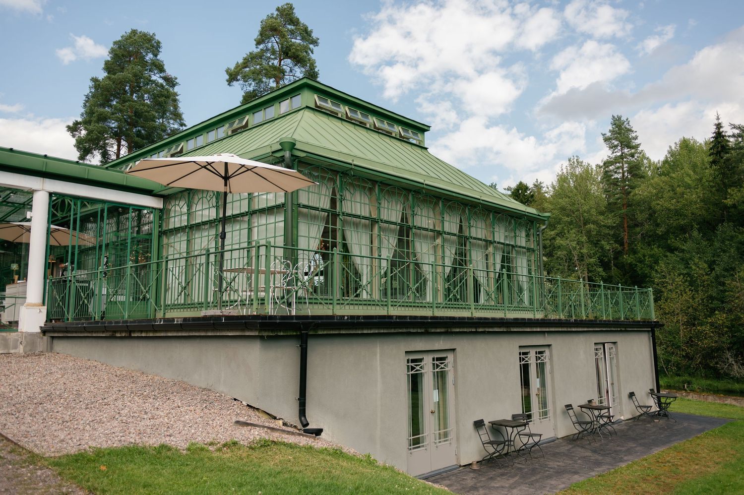 Historic green and white mansion with glass conservatory and landscaped grounds on a sunny day.