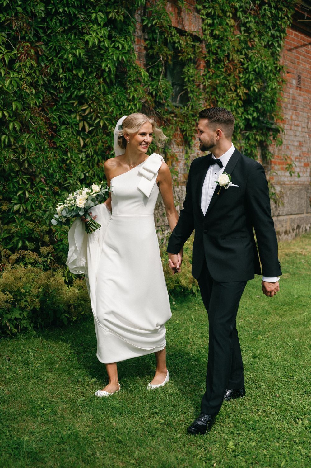 Wedding couple walking hand in hand on grass with ivy-covered brick wall in background.