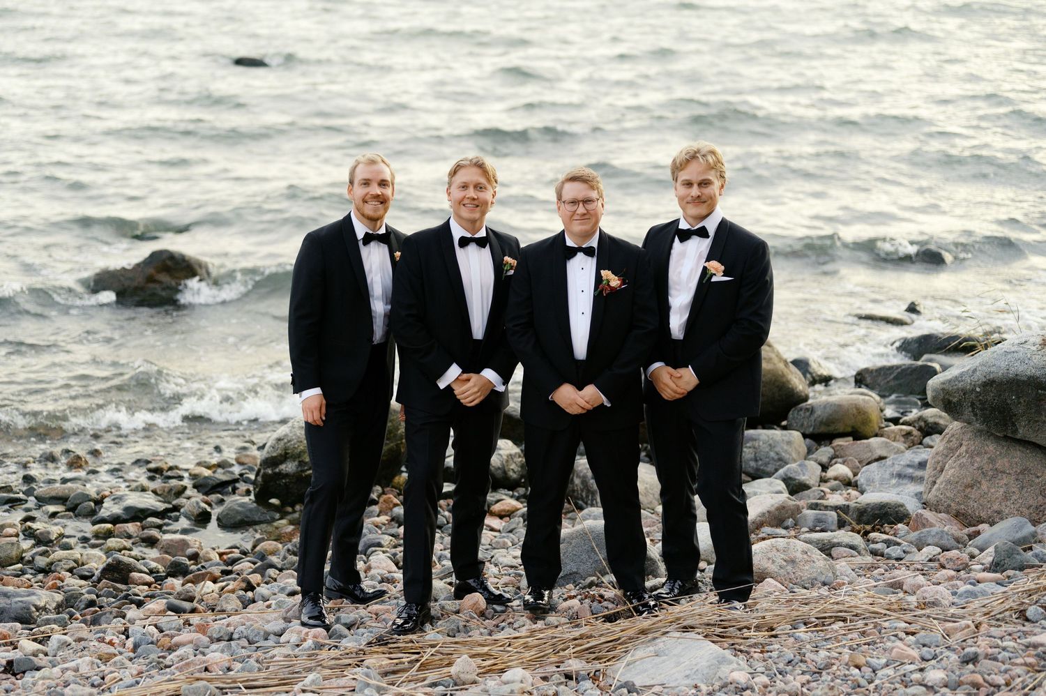 Groomsmen in black tuxedos pose together on a rocky beach with ocean waves in the background.
